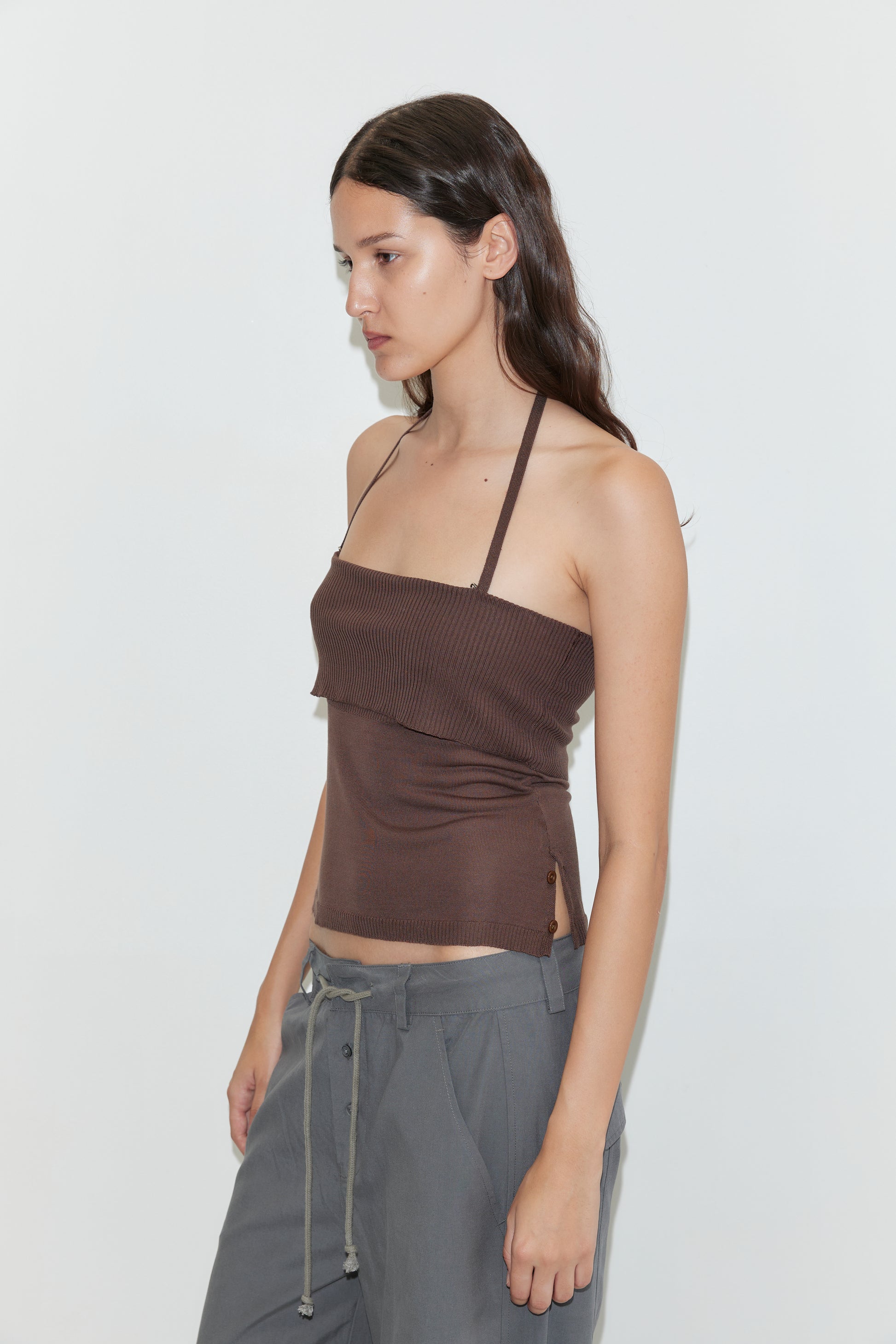 A woman with long brown hair, wearing a brown halter top and Deiji Studios Panel Leg Pant in Charcoal, stands against a plain white background facing left in a relaxed pose.