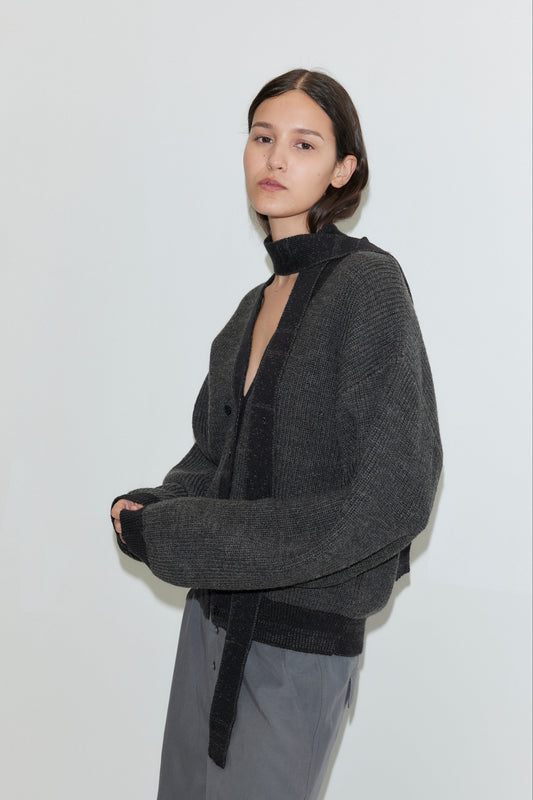 A woman with long dark hair wears the Split Cardigan - Black Contrast, an oversized knit with a deep V-neck, paired with gray pants and standing against a plain white background.