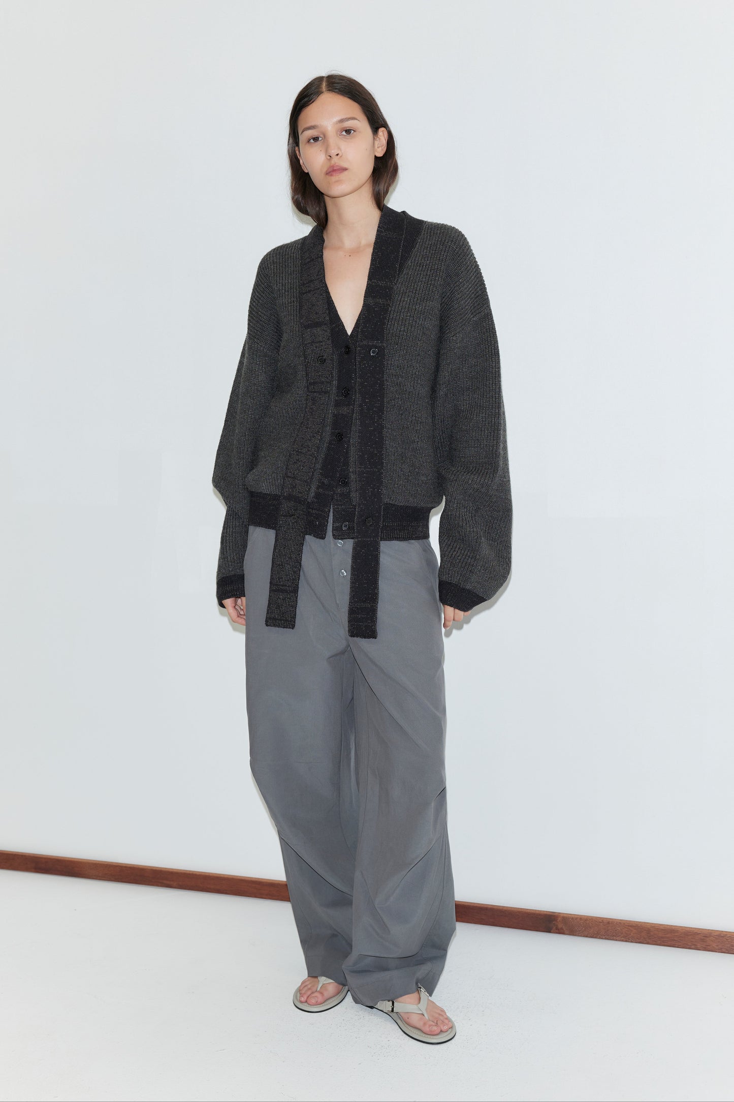 A person with straight, shoulder-length hair wears the Split Cardigan - Black Contrast, featuring a detachable lapel scarf, along with wide gray pants and sandals, standing against a plain white wall with a wooden baseboard.