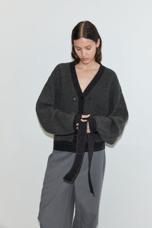 A woman with long brown hair wears the Split Cardigan - Black Contrast, paired with wide gray pants, standing against a plain white background while looking down and holding a fabric belt.
