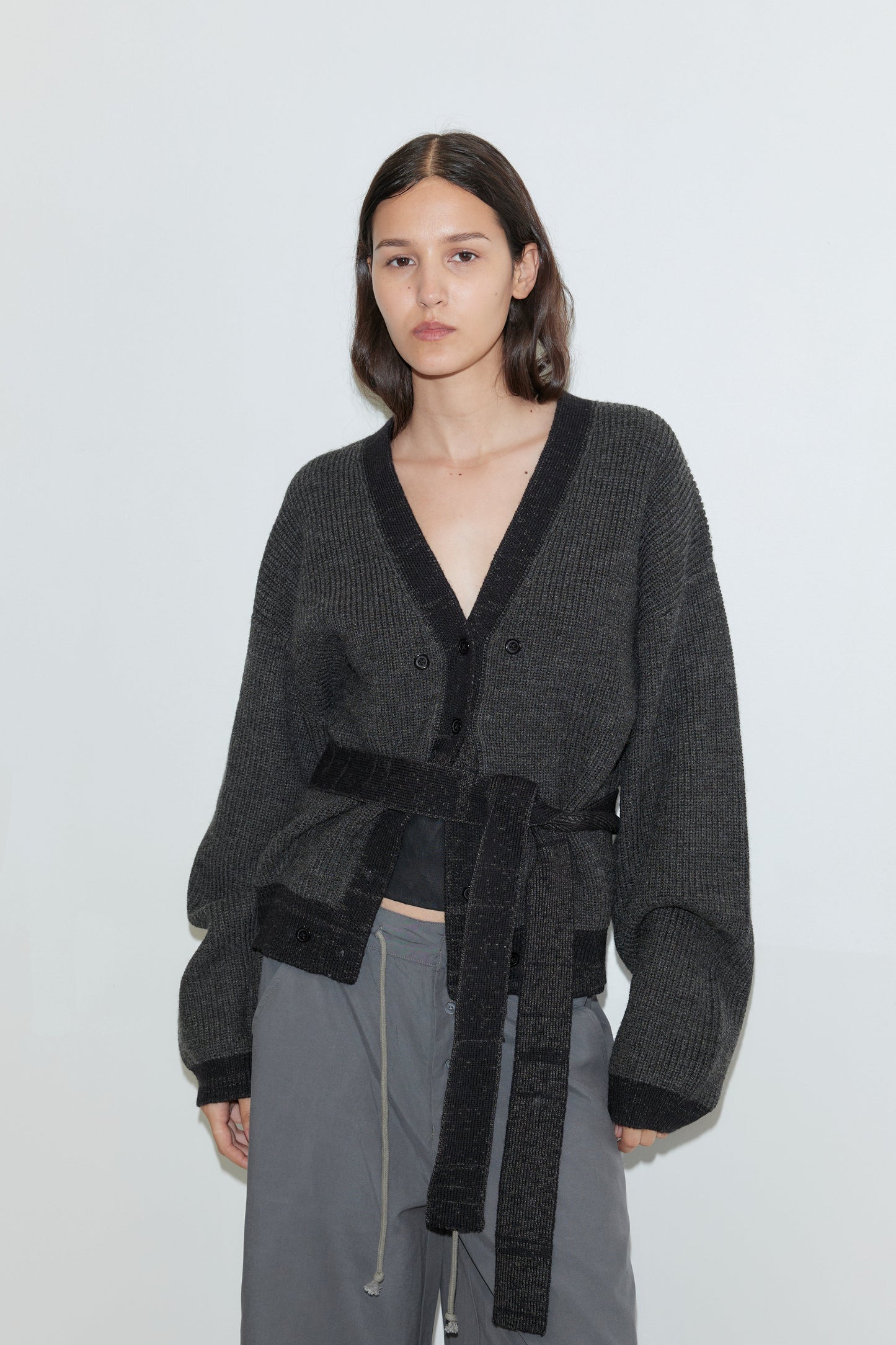 A person with long brown hair wears the Split Cardigan - Black Contrast, an oversized dark gray belted cardigan, paired with gray drawstring pants and stands against a plain light background.