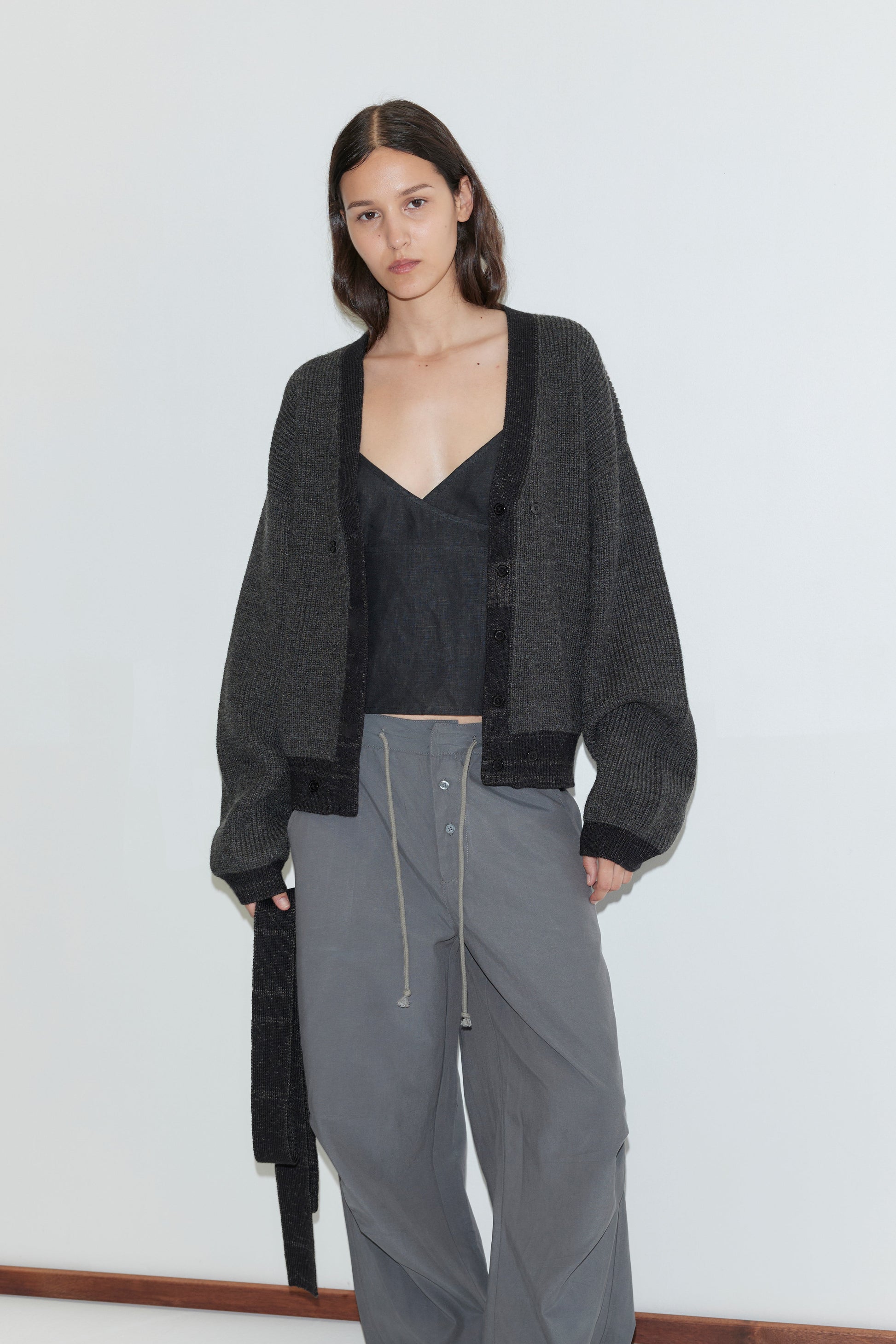 A woman with long dark hair wears the Split Cardigan - Black Contrast, featuring an oversized fit and detachable lapel scarf, over a black top and loose gray drawstring pants, standing against a plain light background.