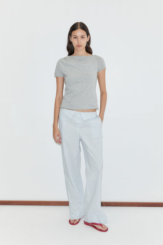 A woman with long dark hair stands against a plain white background, wearing a gray t-shirt, red sandals, and the Collar Point Pant in Mist Stripe. She has one hand in her pocket and a neutral expression.
