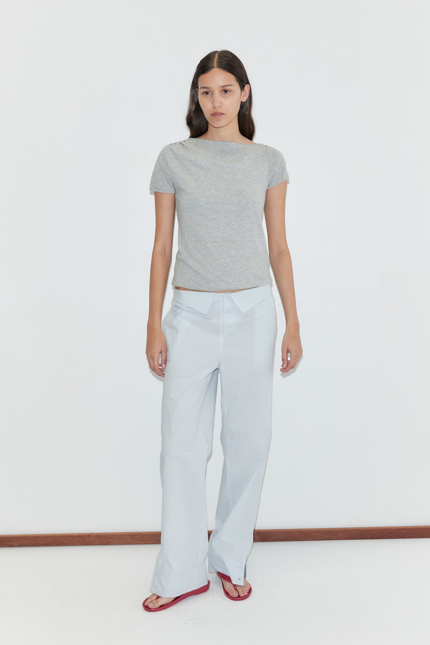 A woman with long dark hair and a neutral expression stands against a plain white background, wearing the Collar Point Pant - Mist Stripe, a fitted grey t-shirt, and red sandals.