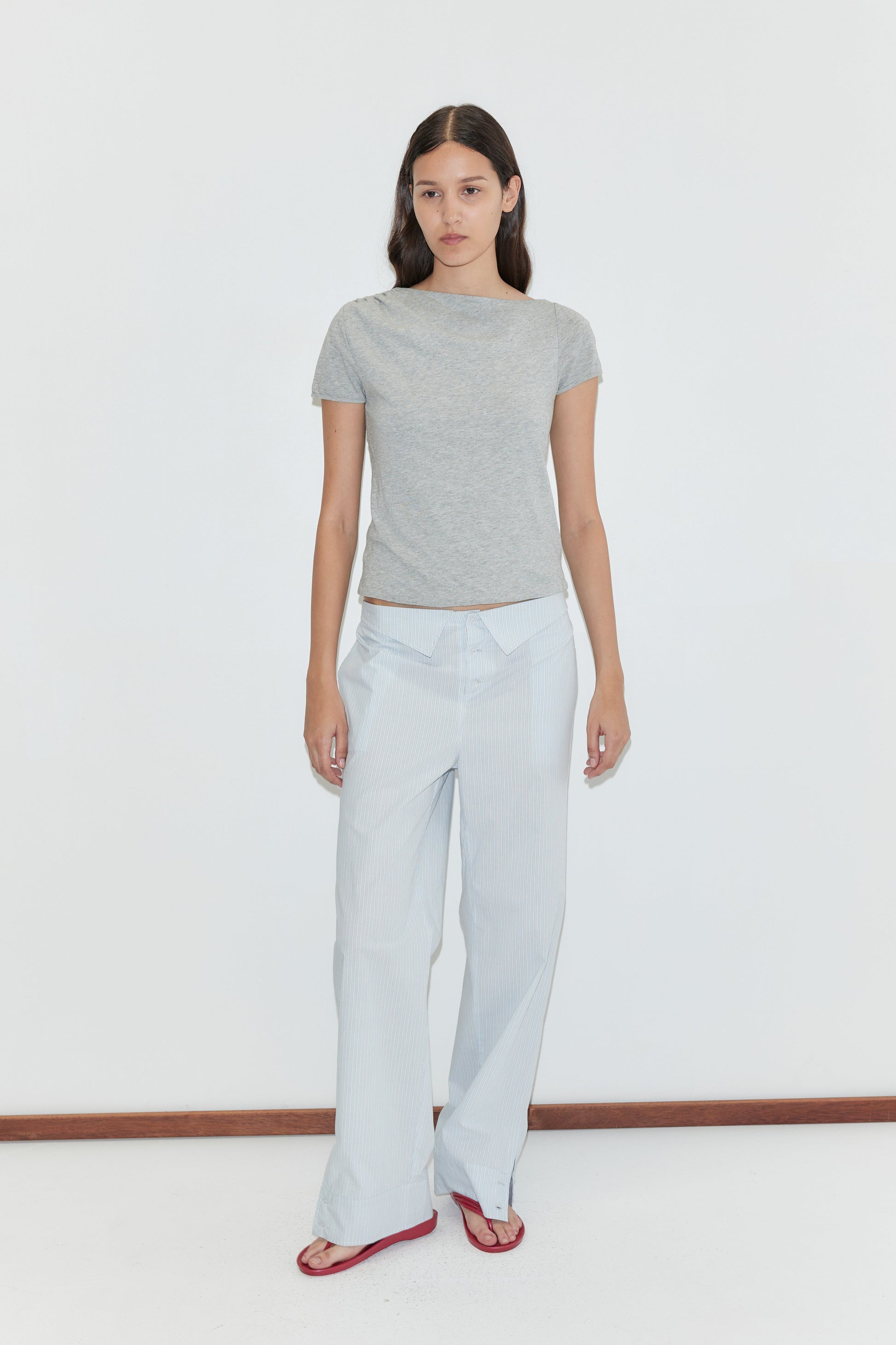 A woman with long dark hair and a neutral expression stands against a plain white background, wearing the Collar Point Pant - Mist Stripe, a fitted grey t-shirt, and red sandals.