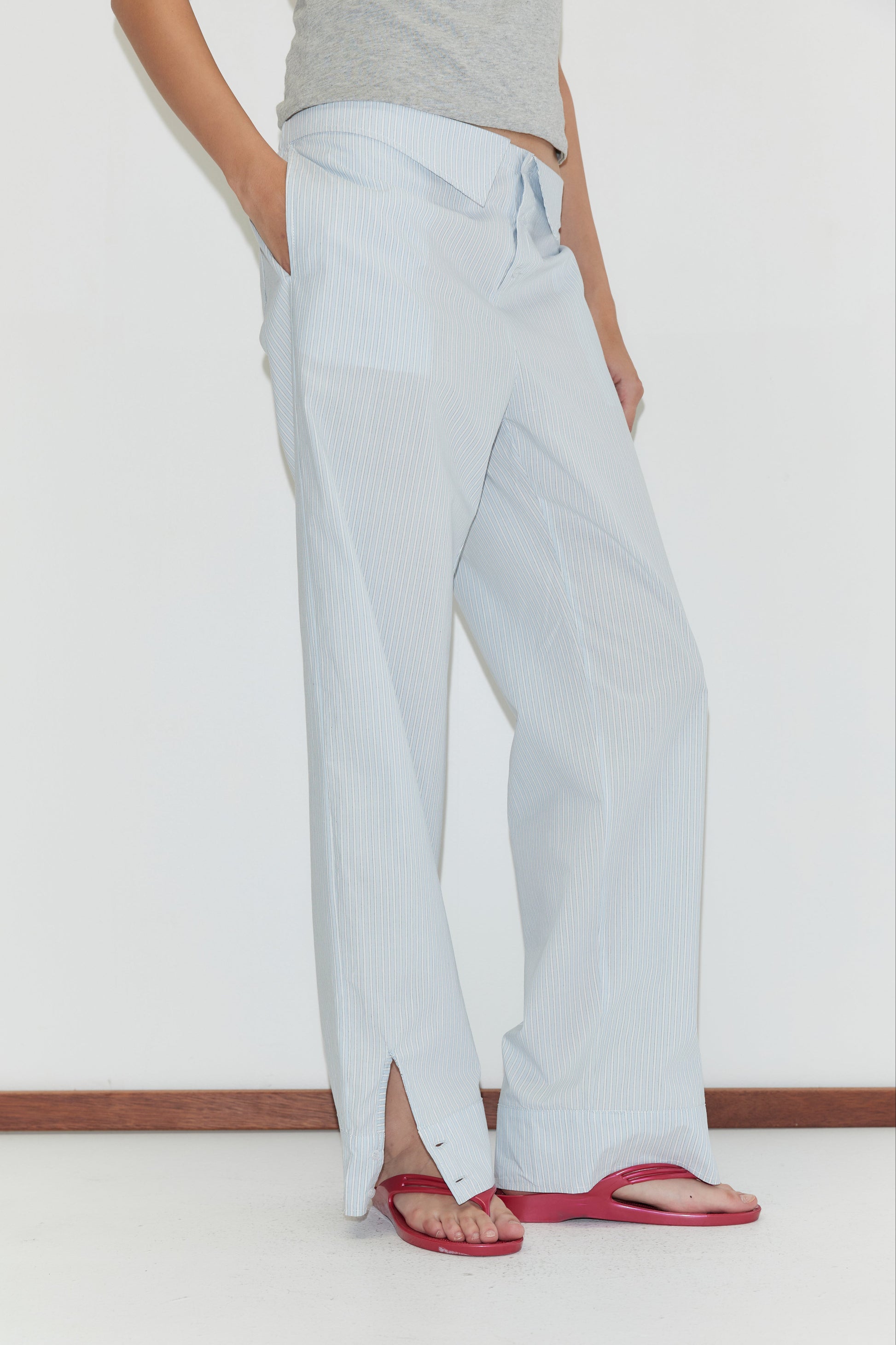 A person stands against a white wall with hands in pockets, wearing the Collar Point Pant - Mist Stripe—light blue pinstripe cotton poplin easy-leg pants with front slits—paired with a gray top and red flip-flops.