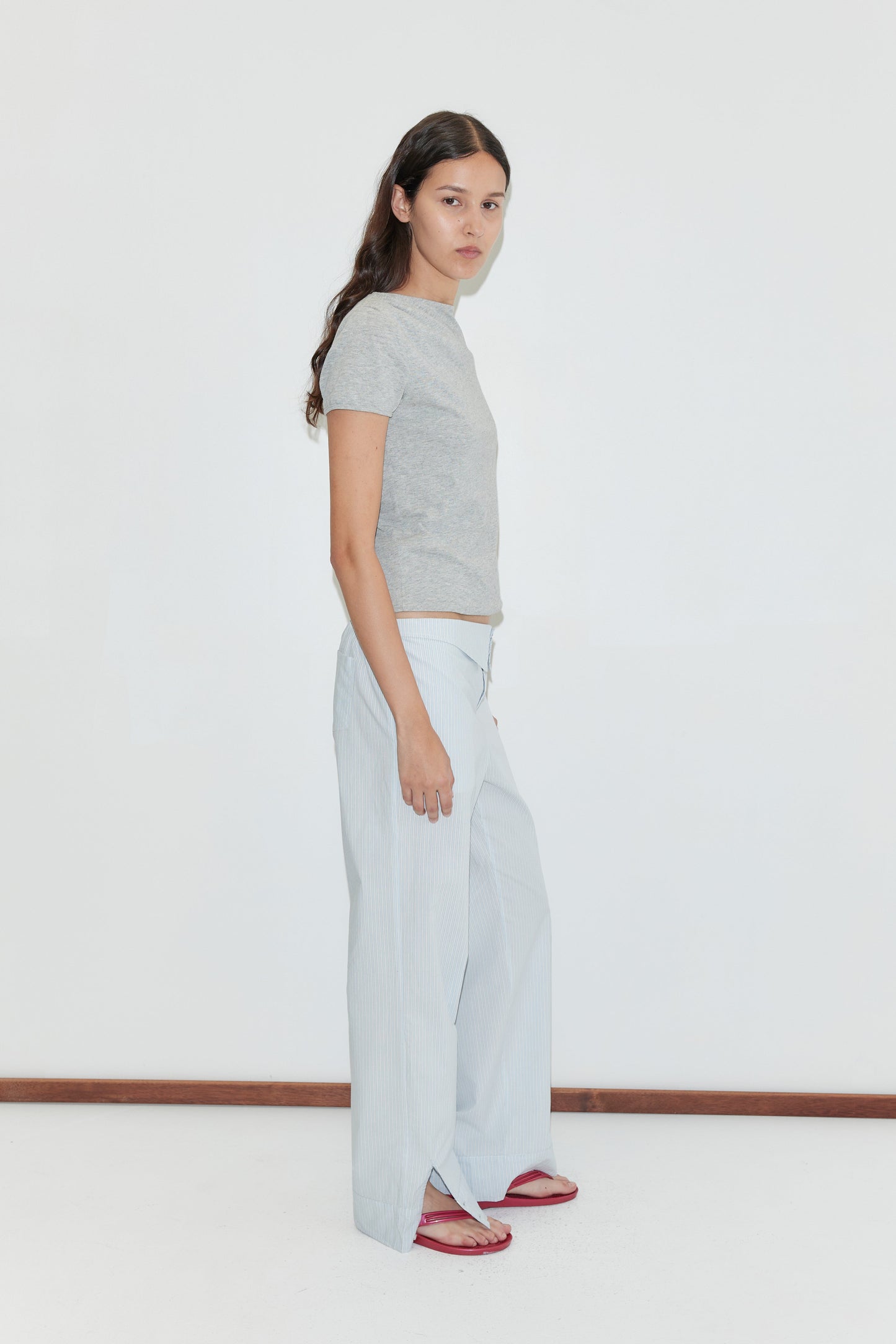 A woman with long dark hair stands sideways against a plain white background, wearing a grey short-sleeve t-shirt, the Collar Point Pant in Mist Stripe, and red sandals.
