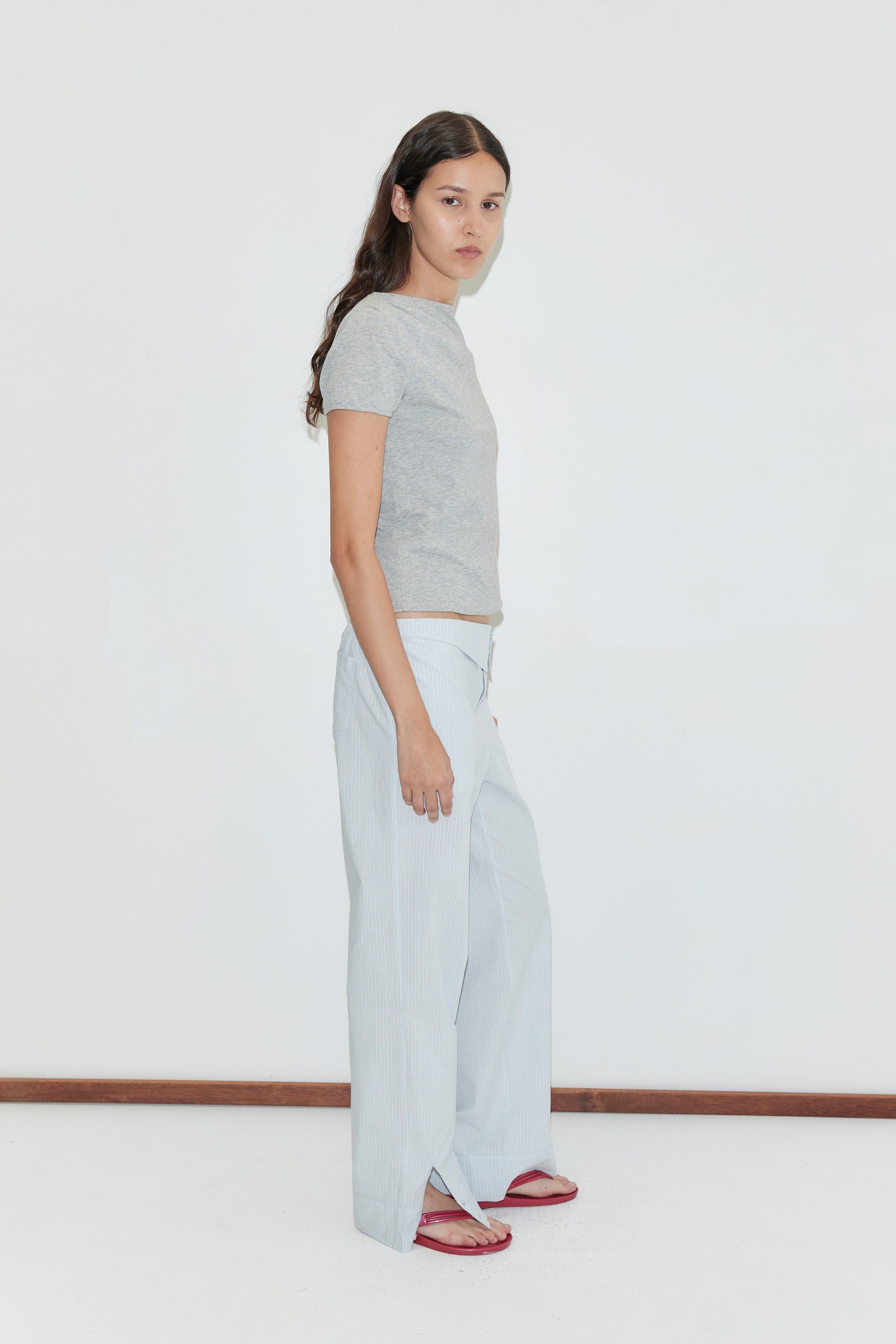 A woman with long dark hair stands sideways against a plain white background, wearing a grey short-sleeve t-shirt, the Collar Point Pant in Mist Stripe, and red sandals.
