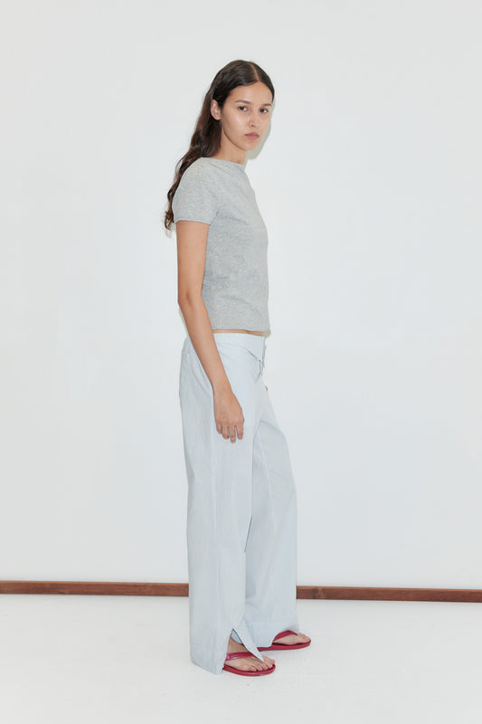 A woman with long dark hair stands sideways against a plain white background, wearing a grey short-sleeve t-shirt, the Collar Point Pant in Mist Stripe, and red sandals.