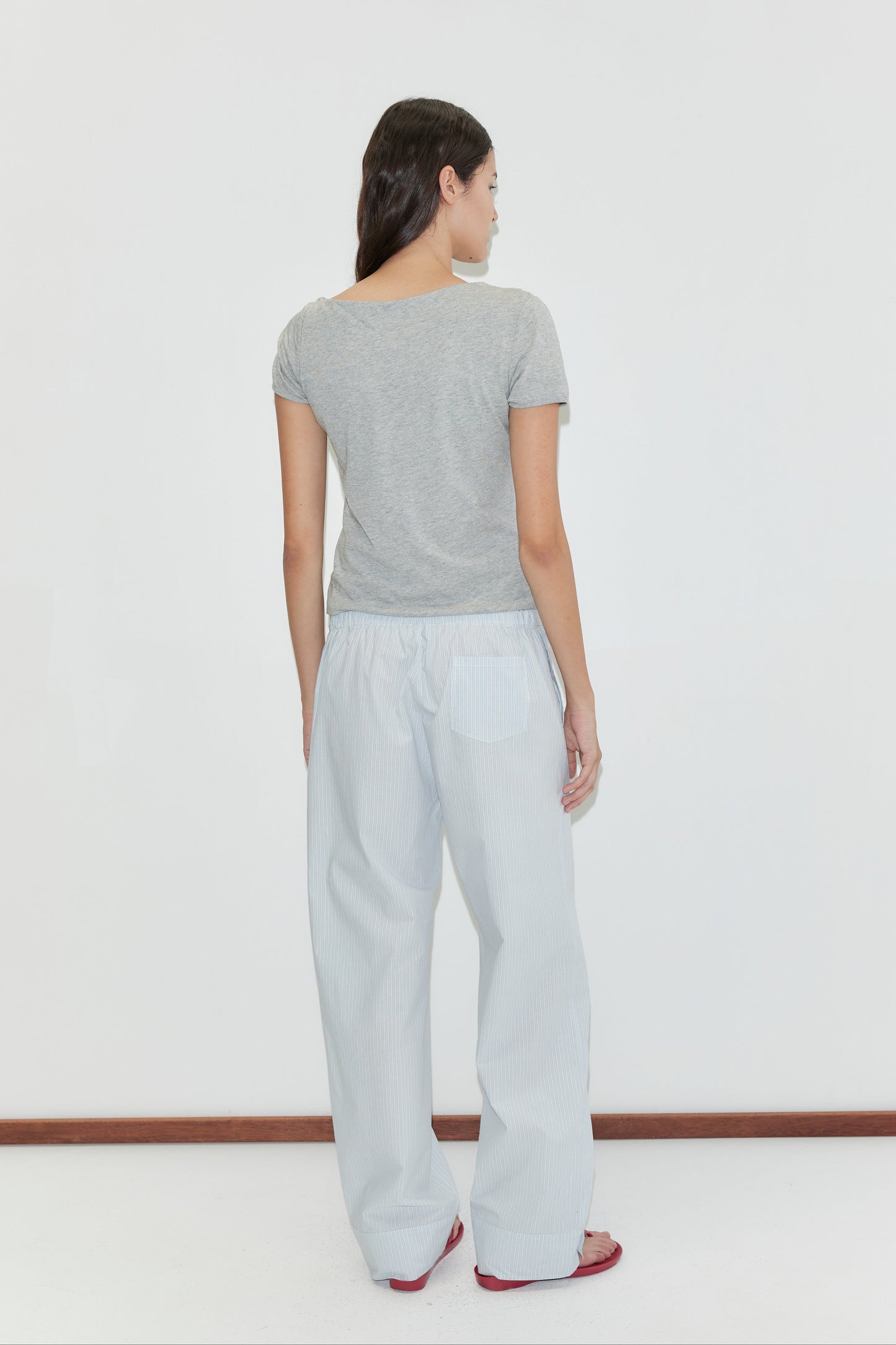 A woman stands facing away against a white background, wearing a short-sleeved gray t-shirt and the Collar Point Pant in Mist Stripe, paired with red sandals.