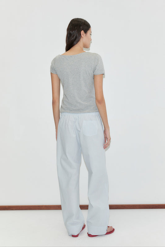 A woman stands facing away against a white background, wearing a short-sleeved gray t-shirt and the Collar Point Pant in Mist Stripe, paired with red sandals.