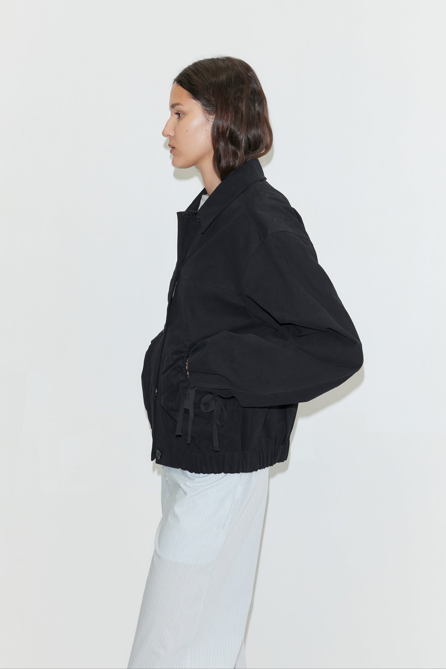 A person with shoulder-length brown hair, wearing the Button Up Jacket - Black and a light-colored skirt, stands in profile against a plain white background.
