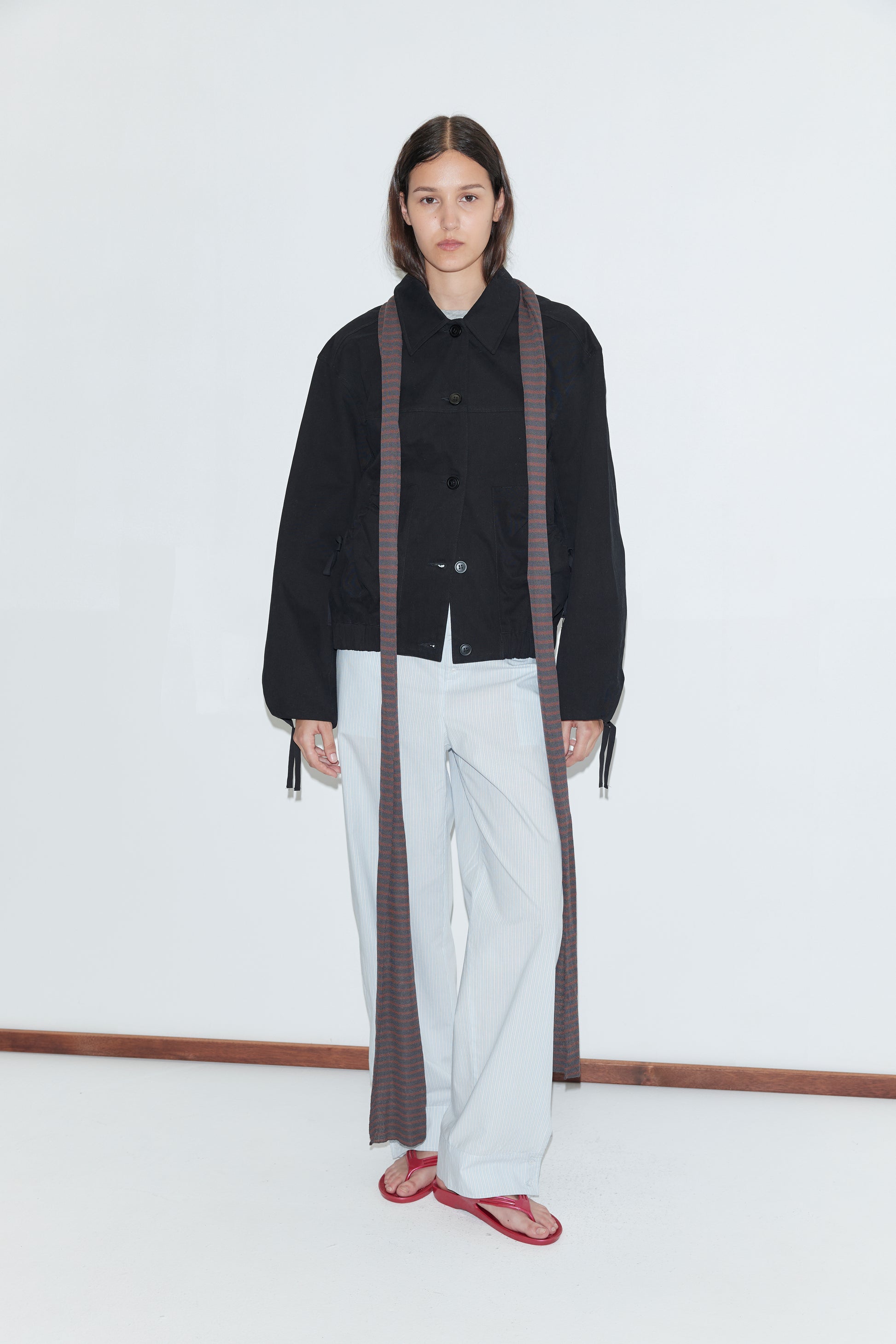 A person with long dark hair and a neutral expression stands against a white background wearing the Button Up Jacket - Black by Deiji Studios, light striped pants, a long scarf draped on both sides, and red sandals.