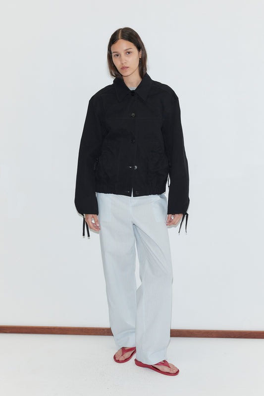 A person stands against a plain white background, wearing the Button Up Jacket - Black from Deiji Studios, loose white striped pants, and red flip-flops. They have straight brown hair and a neutral expression.