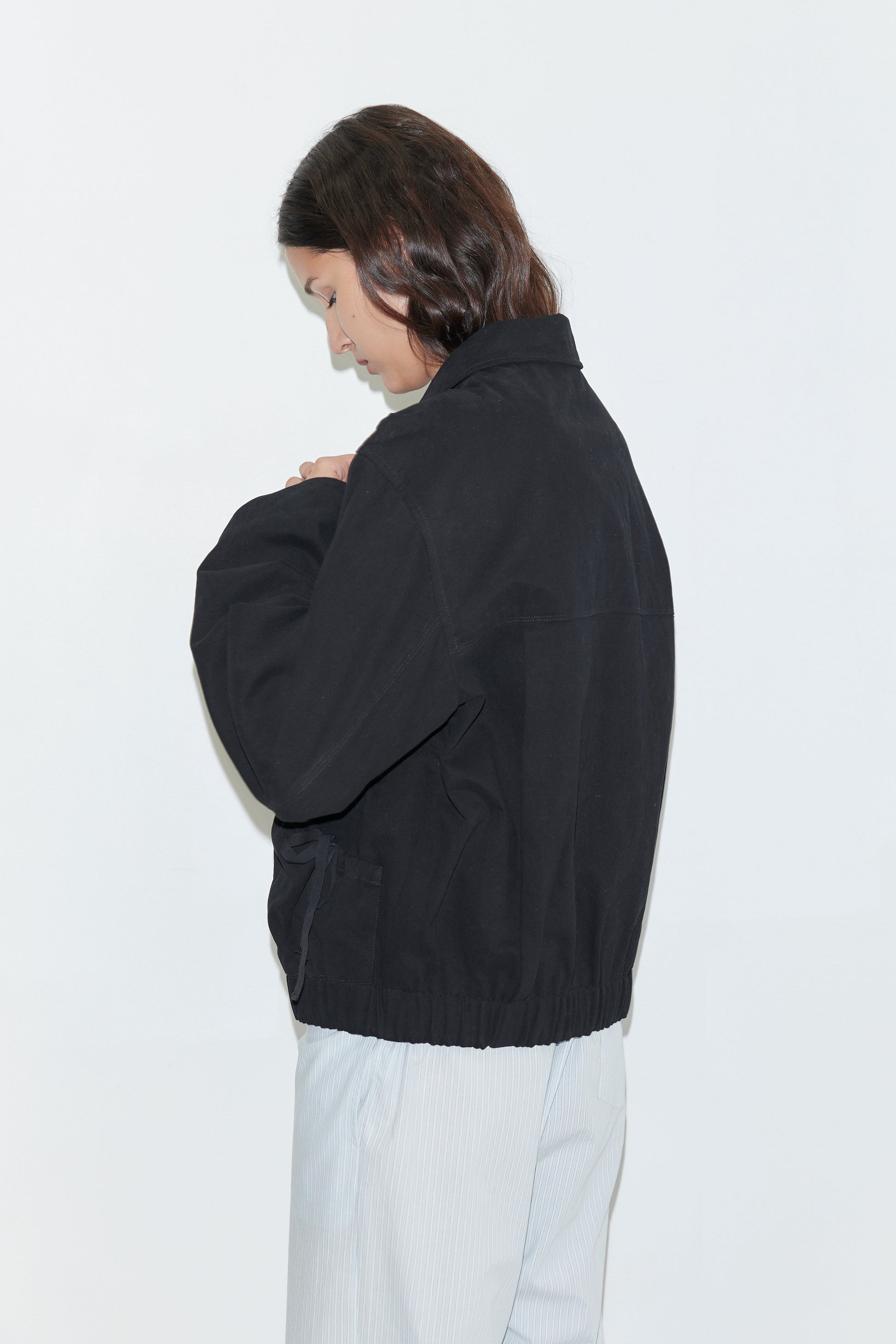 A person with long brown hair wears the Button Up Jacket - Black and light-colored pants, standing sideways against a plain white background, looking down with hands together.