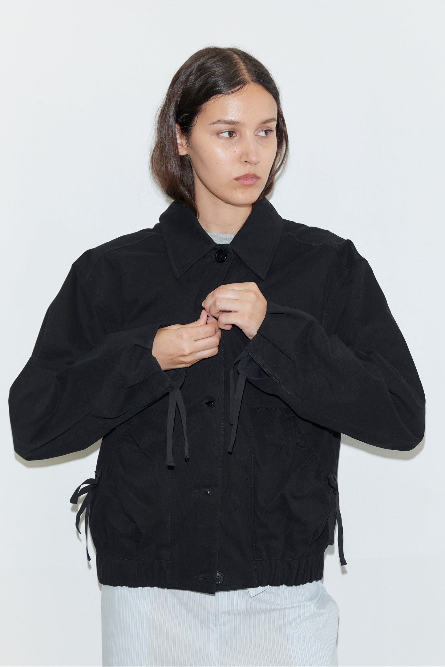 A woman with straight brown hair wears the Button Up Jacket - Black by Deiji Studios, paired with light-colored pants, standing against a plain white background while buttoning her jacket and looking slightly to the side.