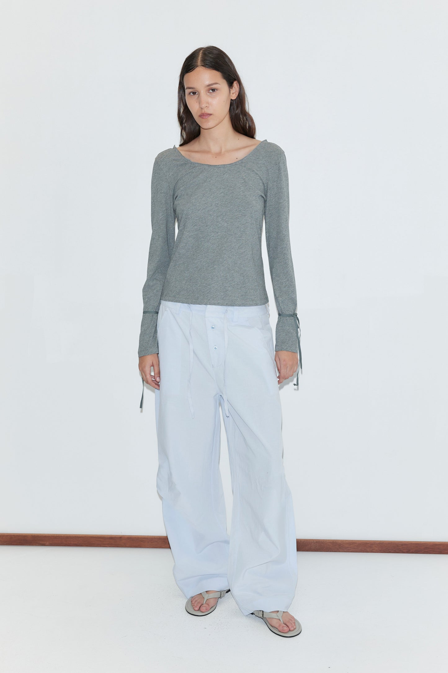 A woman stands against a plain white background, wearing a gray long-sleeve top, the Panel Leg Pant in Powder Blue featuring Deiji Studios' signature relaxed wide-leg silhouette, and sandals. She has straight dark hair and a neutral expression.
