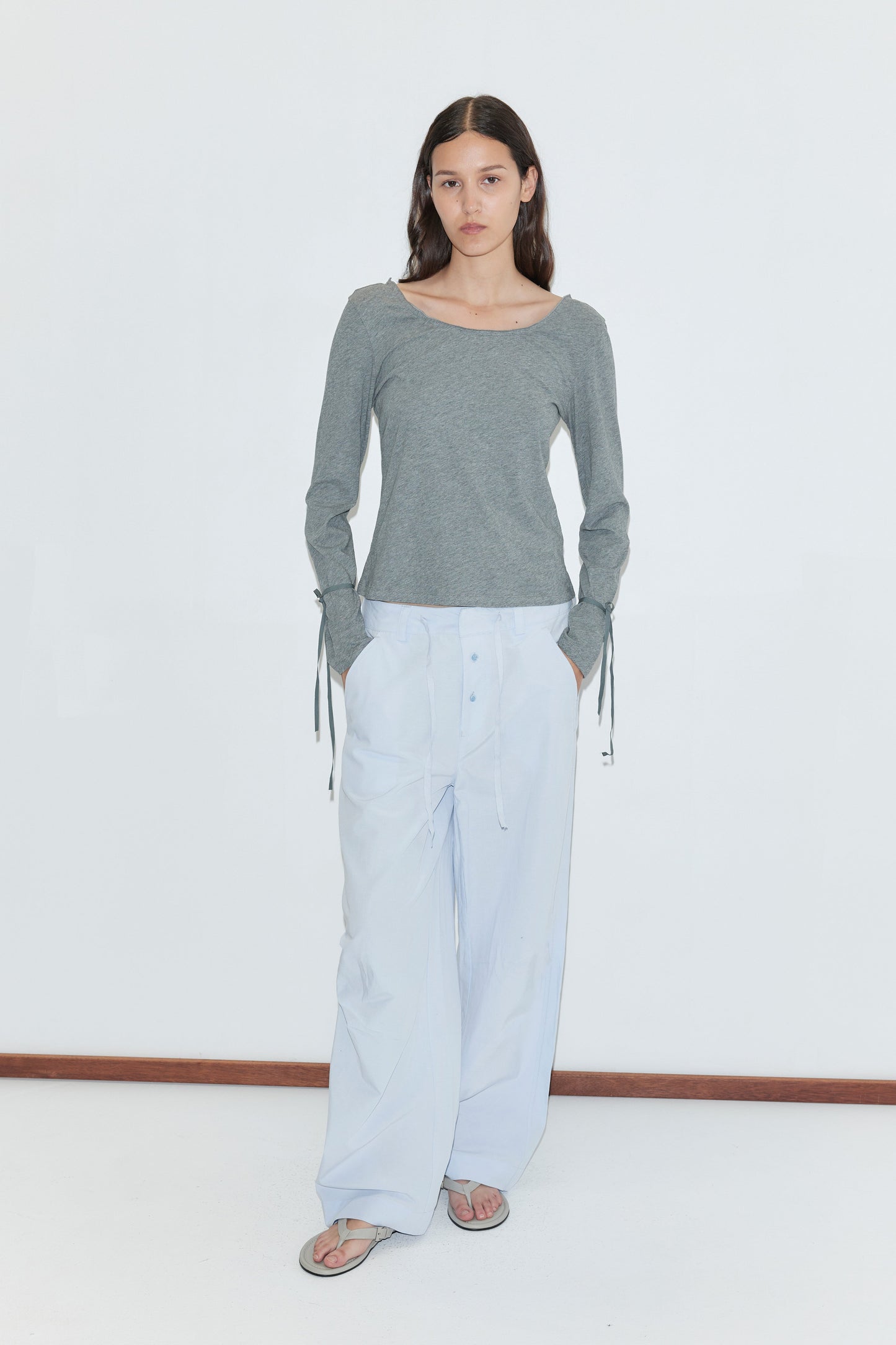 A woman with long brown hair wears a gray long-sleeve shirt with tie details and loose powder blue panel leg pants (Panel Leg Pant - Powder Blue) from Deiji Studios. She stands against a plain white background, looking at the camera, and is wearing sandals.