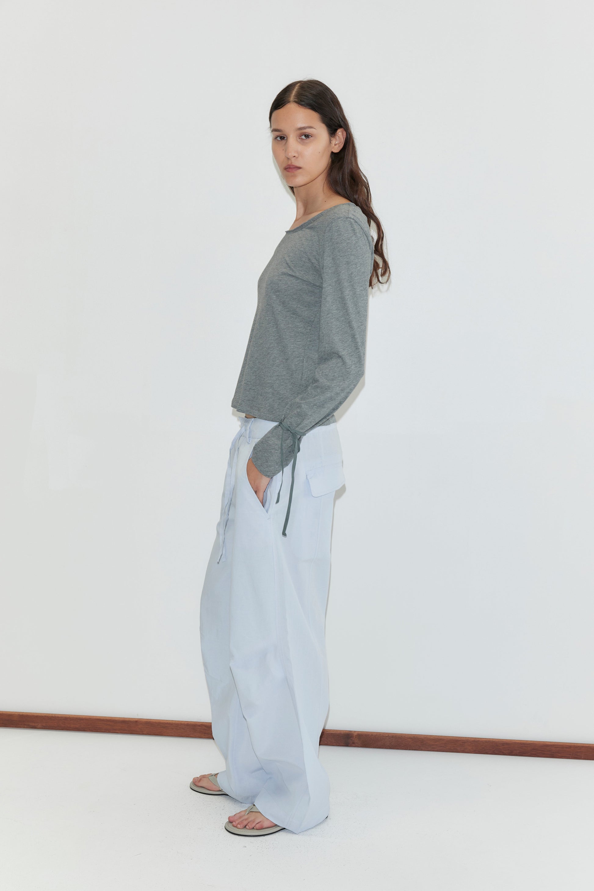 A woman with long brown hair, wearing a gray long-sleeve top, the Panel Leg Pant in Powder Blue, and sandals, stands sideways against a plain white background.