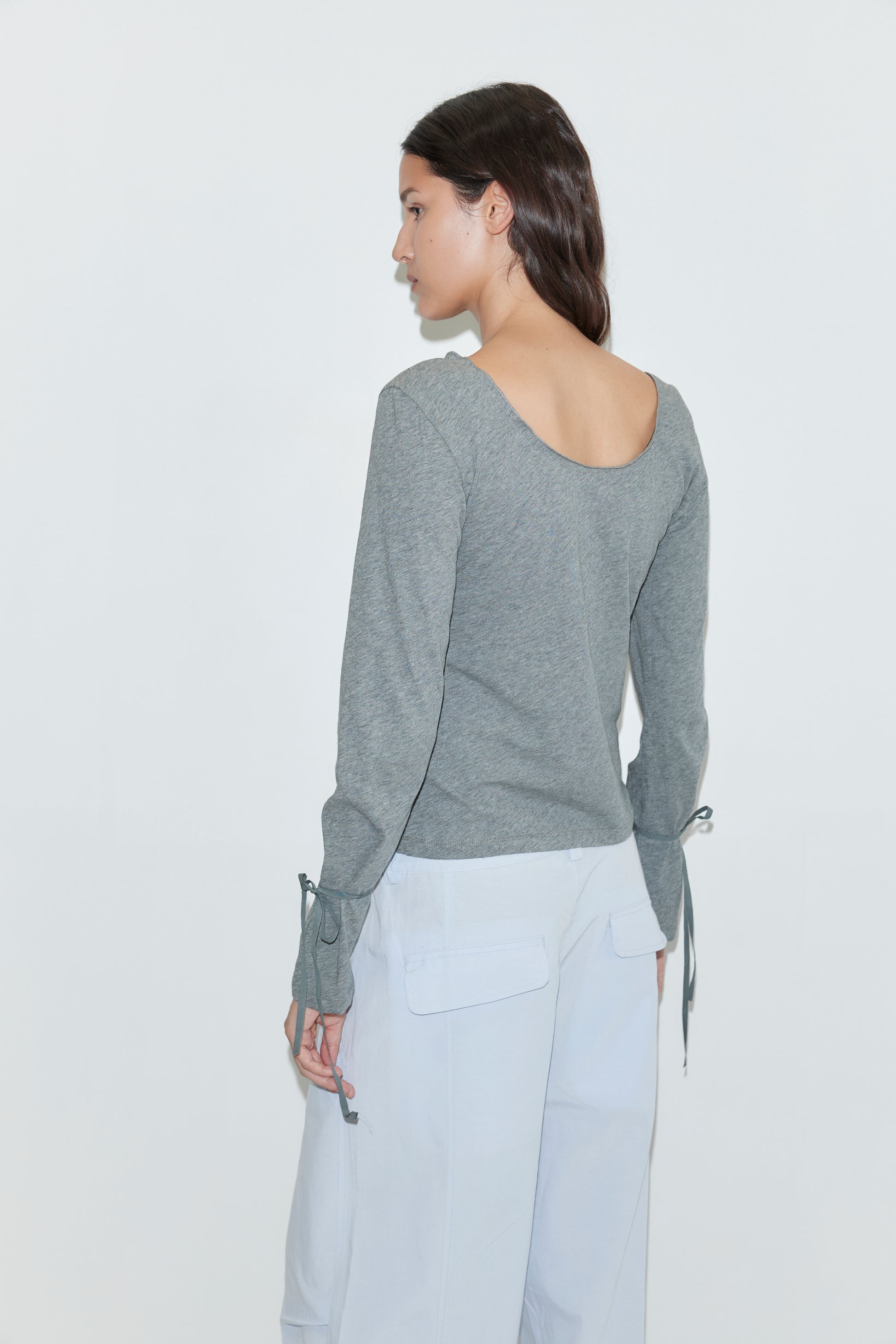 A woman with long dark hair stands sideways, looking left. She wears a gray long-sleeve top with tied wrists and Deiji Studios' Panel Leg Pant in powder blue. The background is plain white.