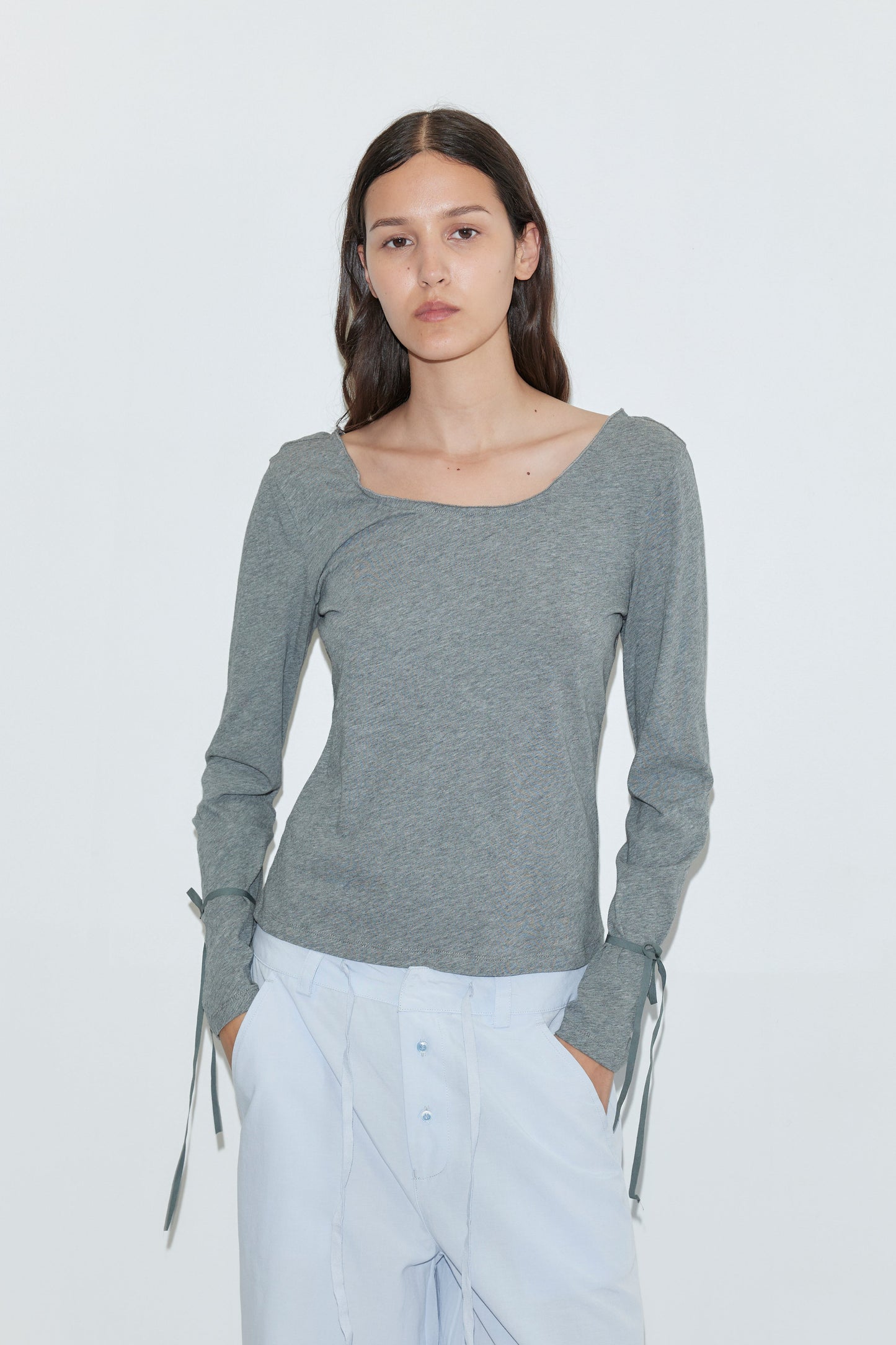 A young woman with long dark hair wears the Deiji Studios Cuff Tie Top in grey and light blue pants, standing against a plain white background and looking directly at the camera with a neutral expression.