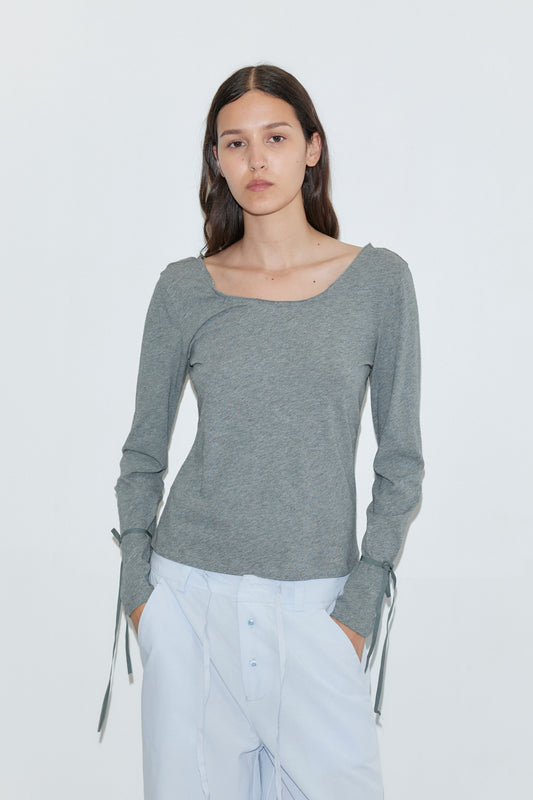 A young woman with long dark hair wears the Deiji Studios Cuff Tie Top in grey and light blue pants, standing against a plain white background and looking directly at the camera with a neutral expression.