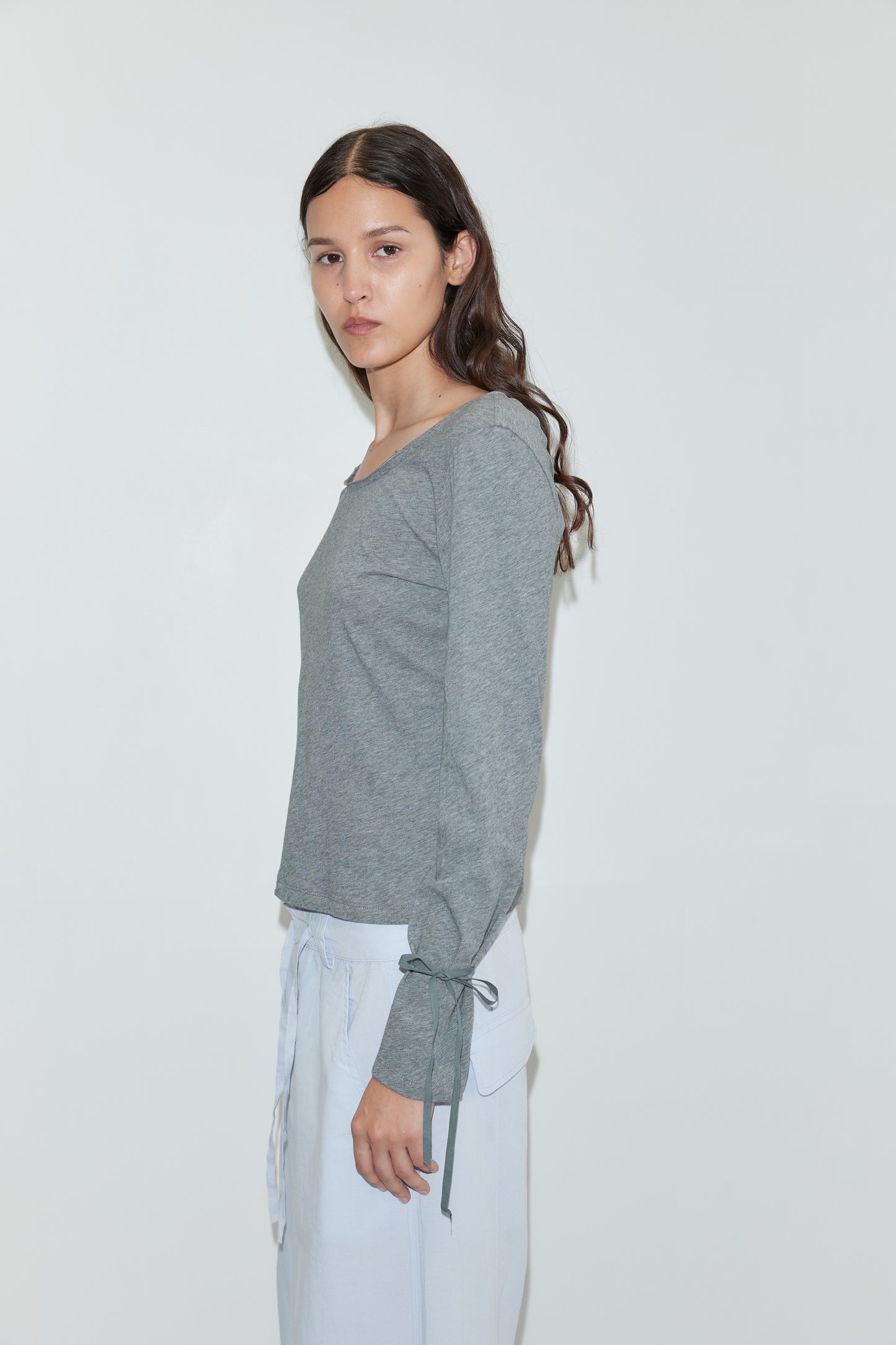 A young woman with long, wavy brown hair stands sideways against a white background, wearing the Cuff Tie Top - Grey by Deiji Studios with ribbon detail at the wrist and light-colored pants, calmly looking toward the camera.