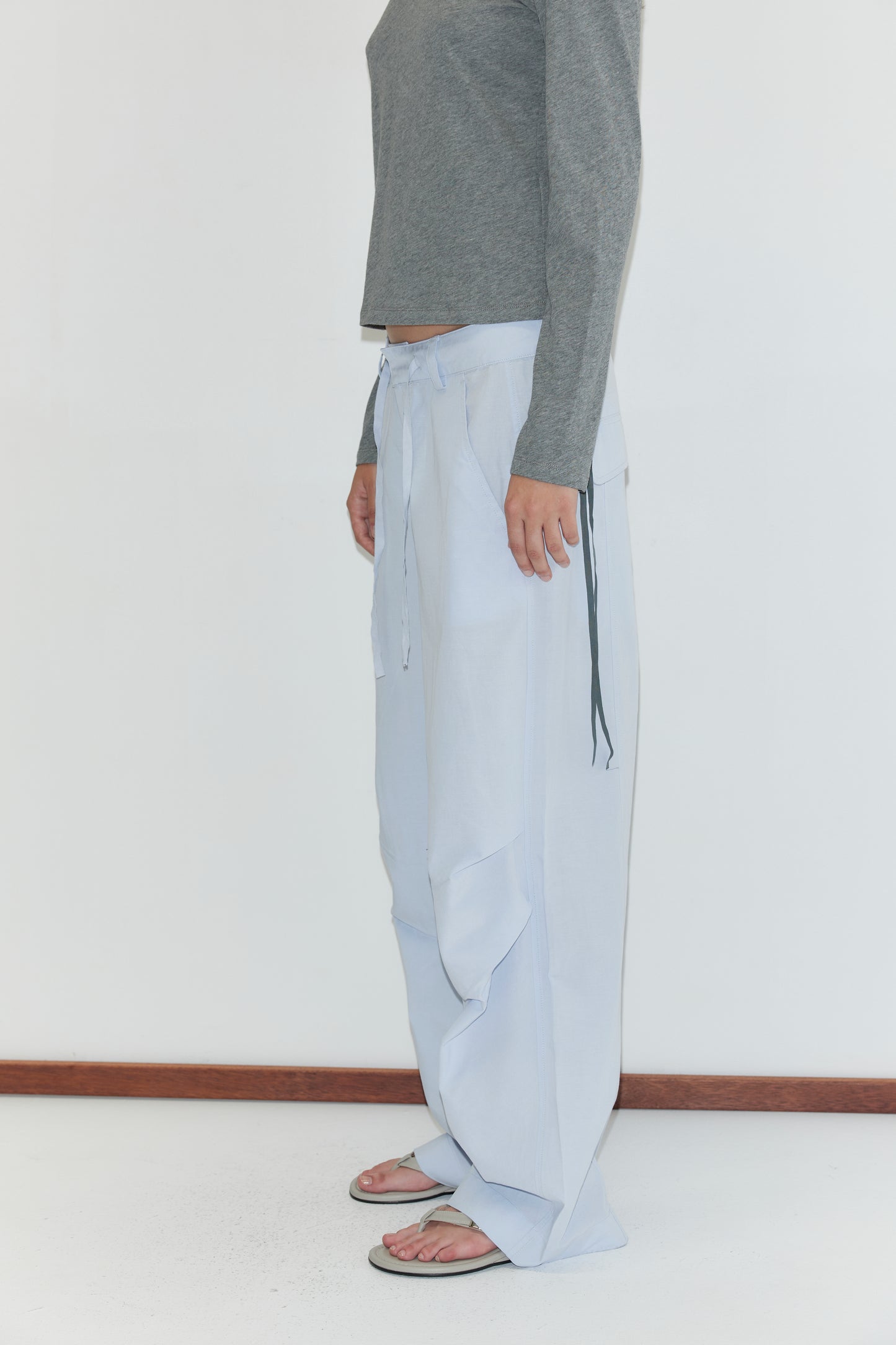 A person stands sideways against a white background, wearing a grey long-sleeve top, the Panel Leg Pant - Powder Blue by Deiji Studios with tie details, and black flip-flops. Only their body from the shoulders down is visible.