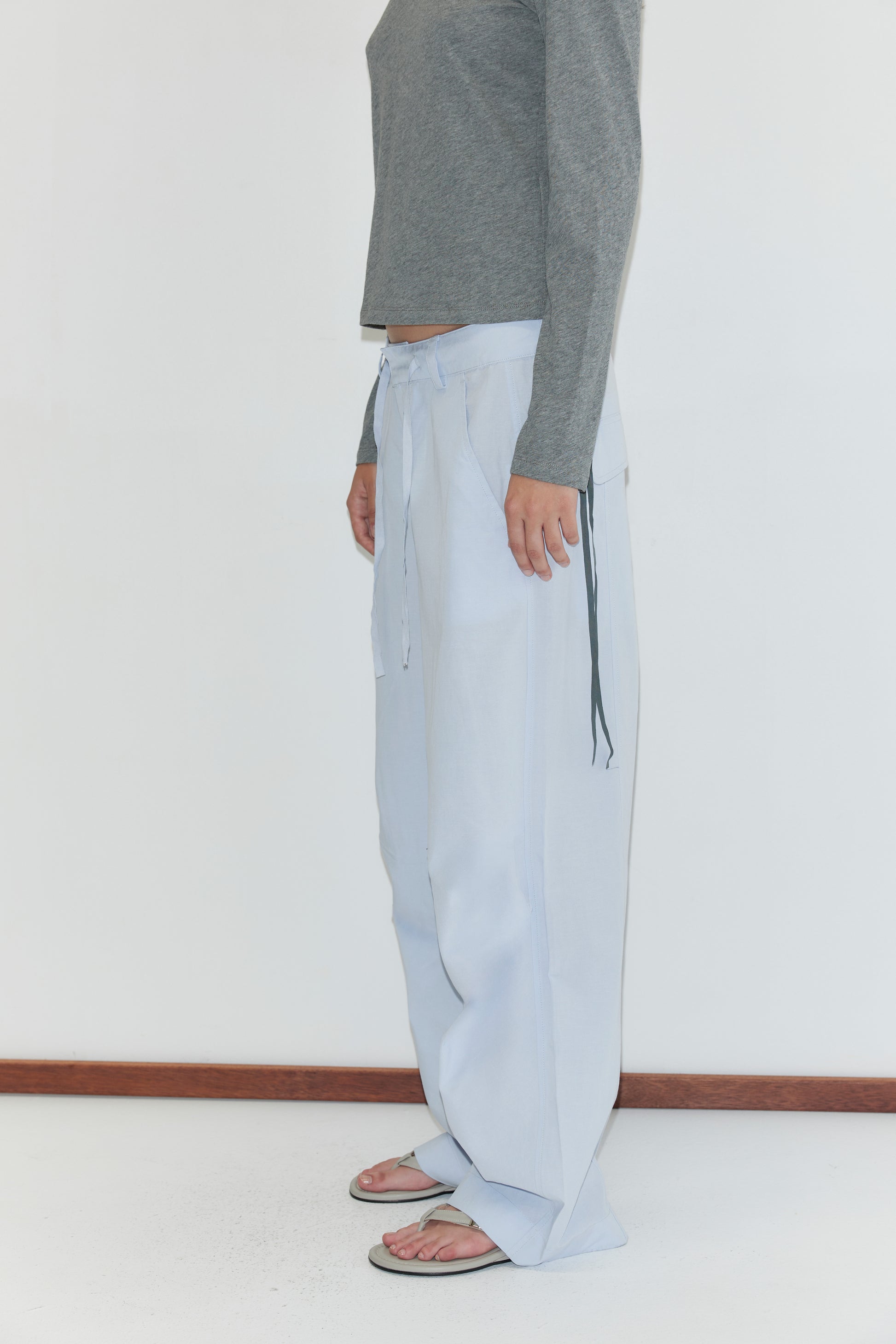 A person stands sideways wearing a gray long-sleeve shirt, the Panel Leg Pant - Powder Blue with a drawstring, and sandals. The background is plain white, and their face is not visible.