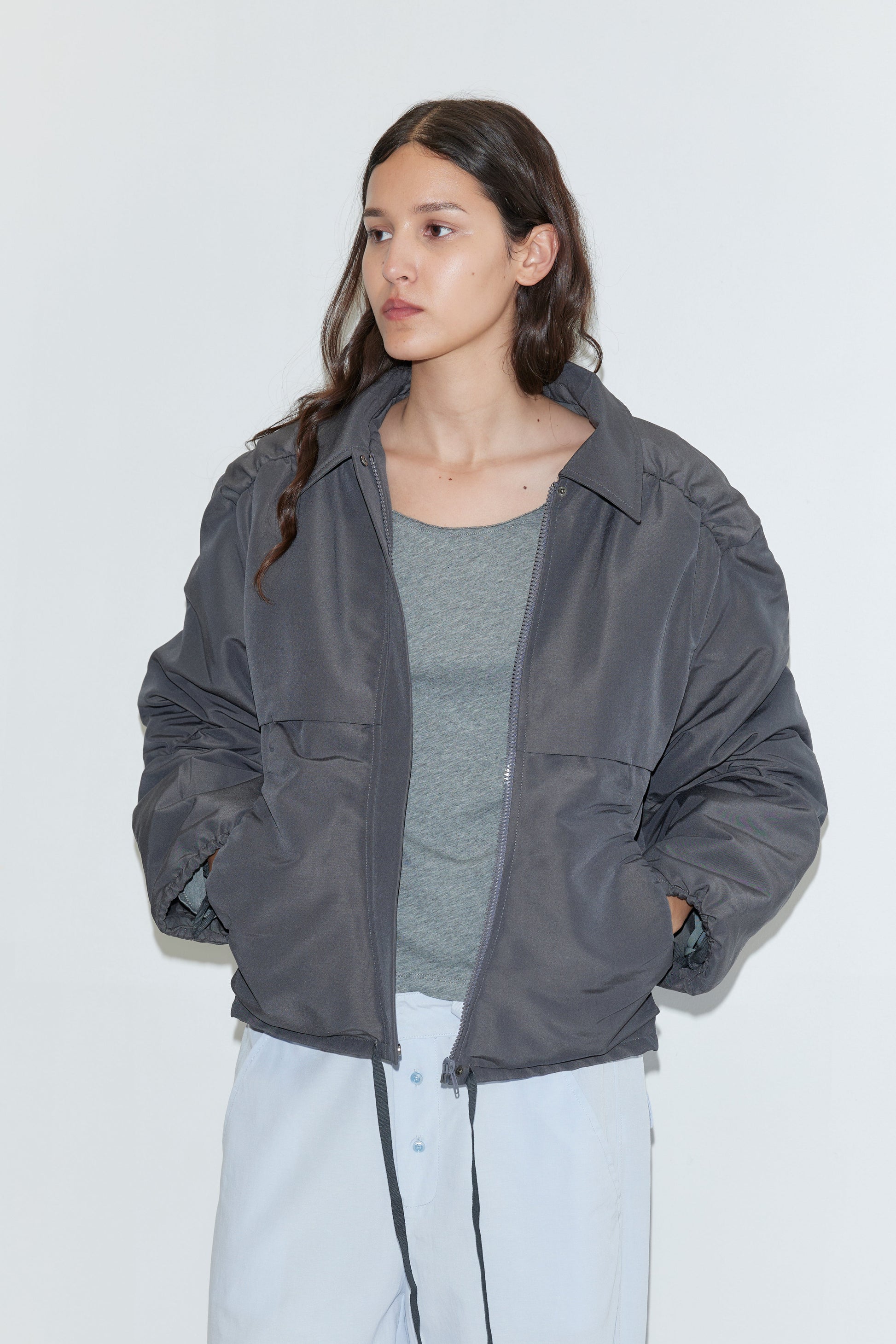 A woman with long brown hair stands against a plain white background, wearing the Gathered Jacket - Slate by Deiji Studios over a gray top and light blue pants, with her hands in her jacket pockets.