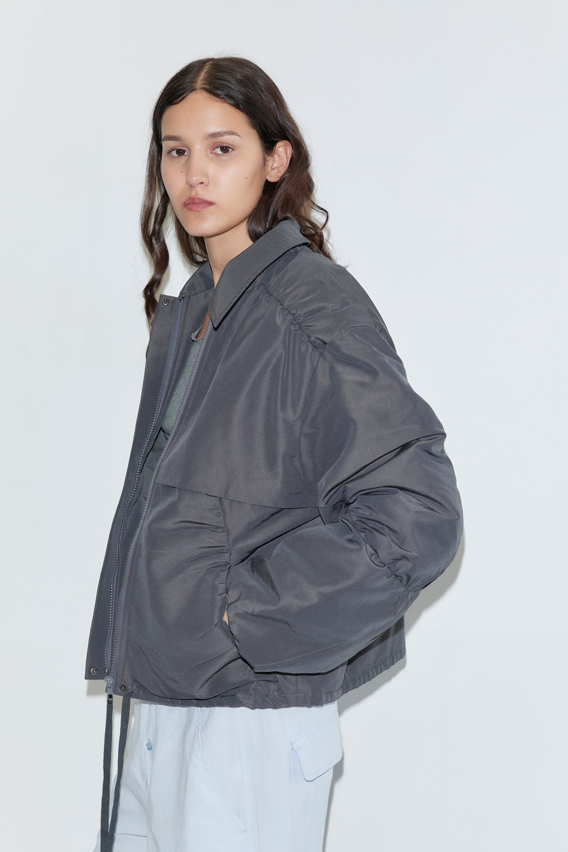 A person with long brown hair wears the Deiji Studios Gathered Jacket - Slate over a light shirt and pants, standing against a plain white background with a neutral expression and one hand in their pocket.
