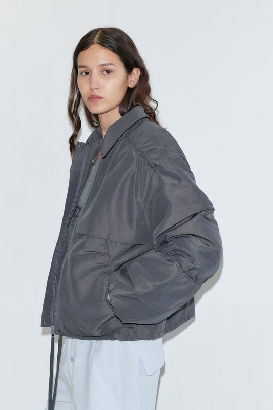 A person with long brown hair wears the Deiji Studios Gathered Jacket - Slate over a light shirt and pants, standing against a plain white background with a neutral expression and one hand in their pocket.