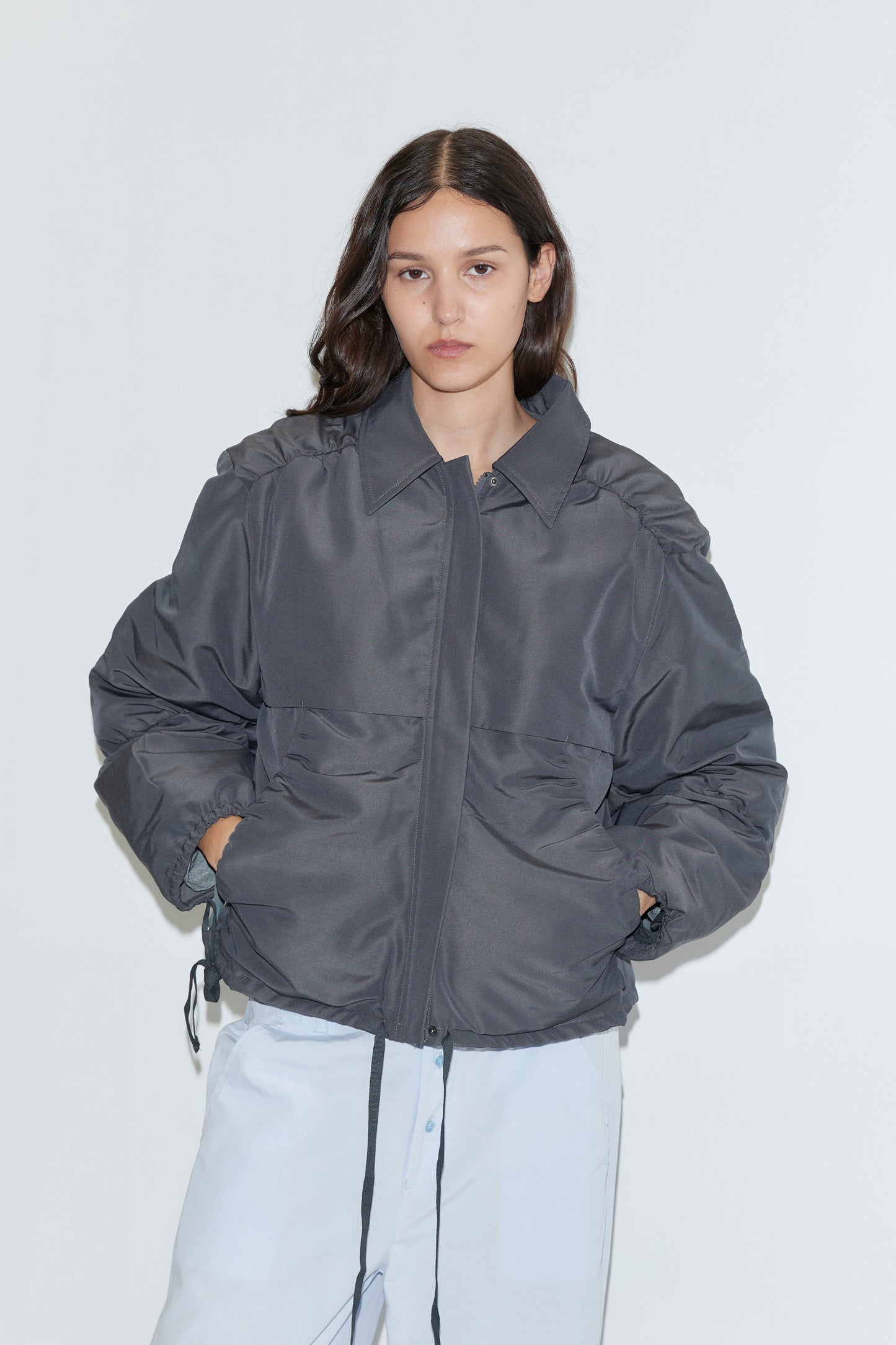 A person with long dark hair wears the Gathered Jacket - Slate from Deiji Studios, an oversized lightweight jacket with front pockets and drawstrings, paired with light blue pants against a plain white background.