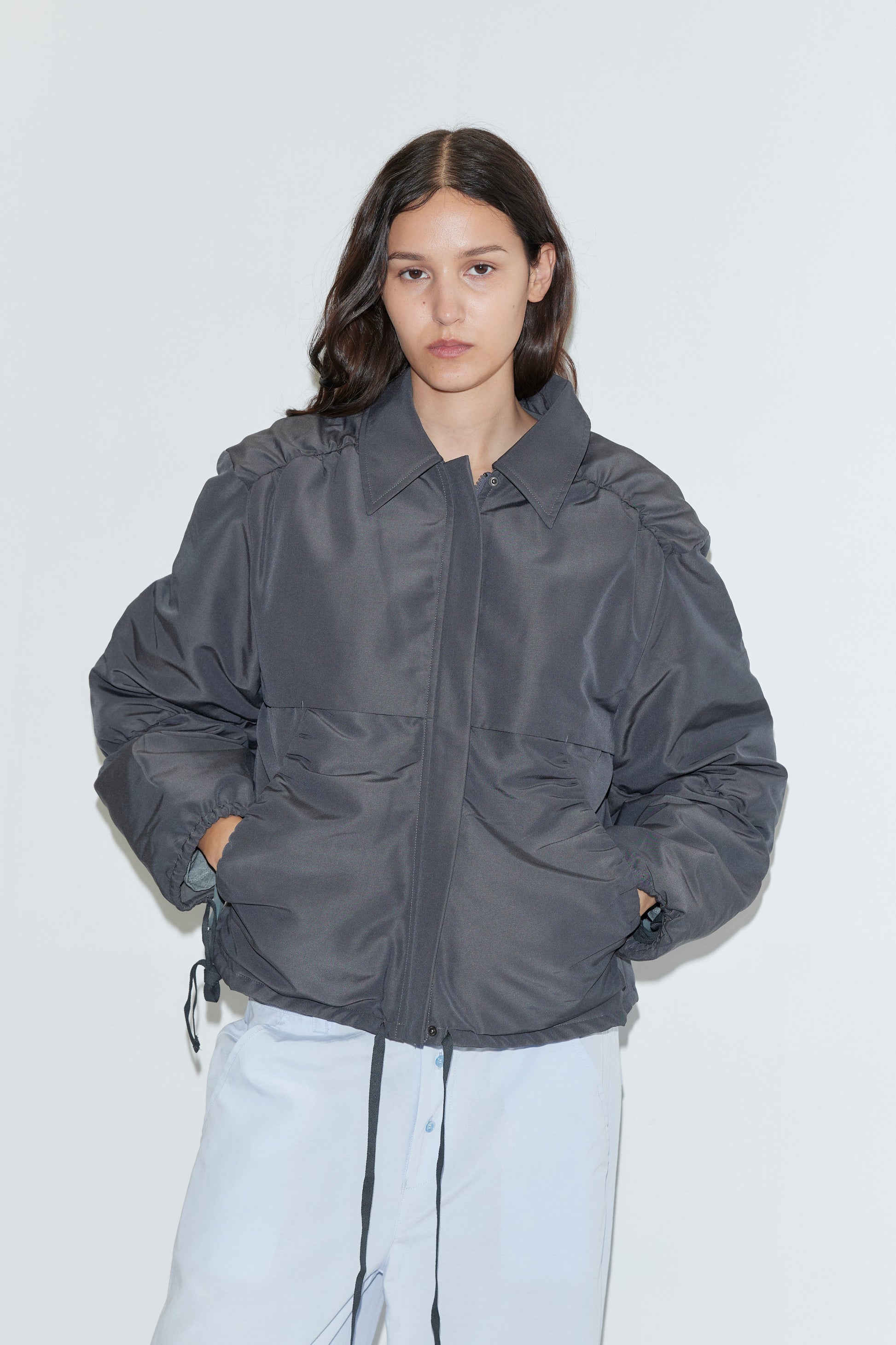 A person with long dark hair wears the Gathered Jacket - Slate from Deiji Studios, an oversized lightweight jacket with front pockets and drawstrings, paired with light blue pants against a plain white background.