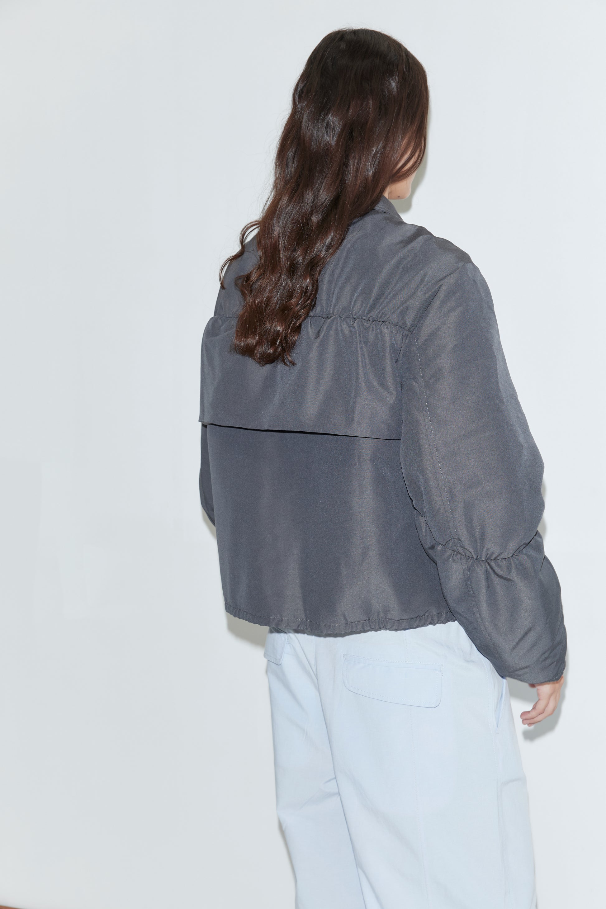 A person with long brown wavy hair, wearing the Deiji Studios Gathered Jacket - Slate and light blue pants, stands with their back to the camera facing a white wall.