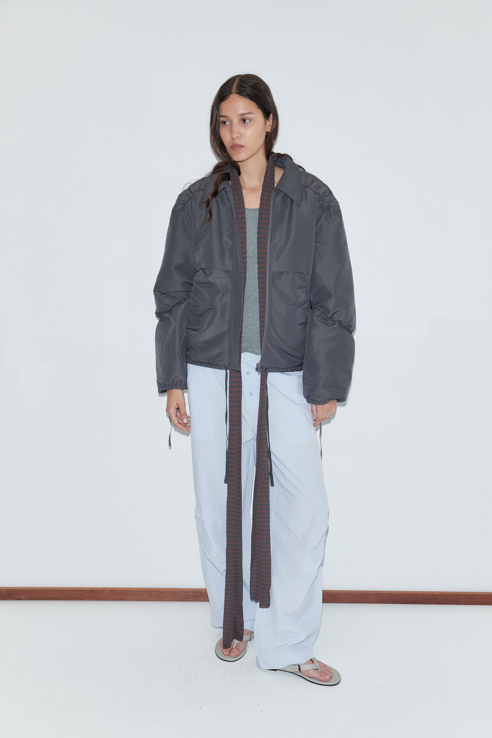 A person with long dark hair wears the Gathered Jacket - Slate by Deiji Studios over a gray top, paired with baggy light blue pants with long ties and flip-flops, standing against a plain white wall and wooden floor.