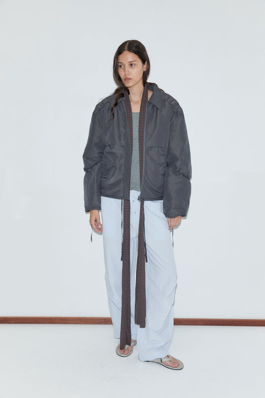 A person with long dark hair wears the Gathered Jacket - Slate by Deiji Studios over a gray top, paired with baggy light blue pants with long ties and flip-flops, standing against a plain white wall and wooden floor.