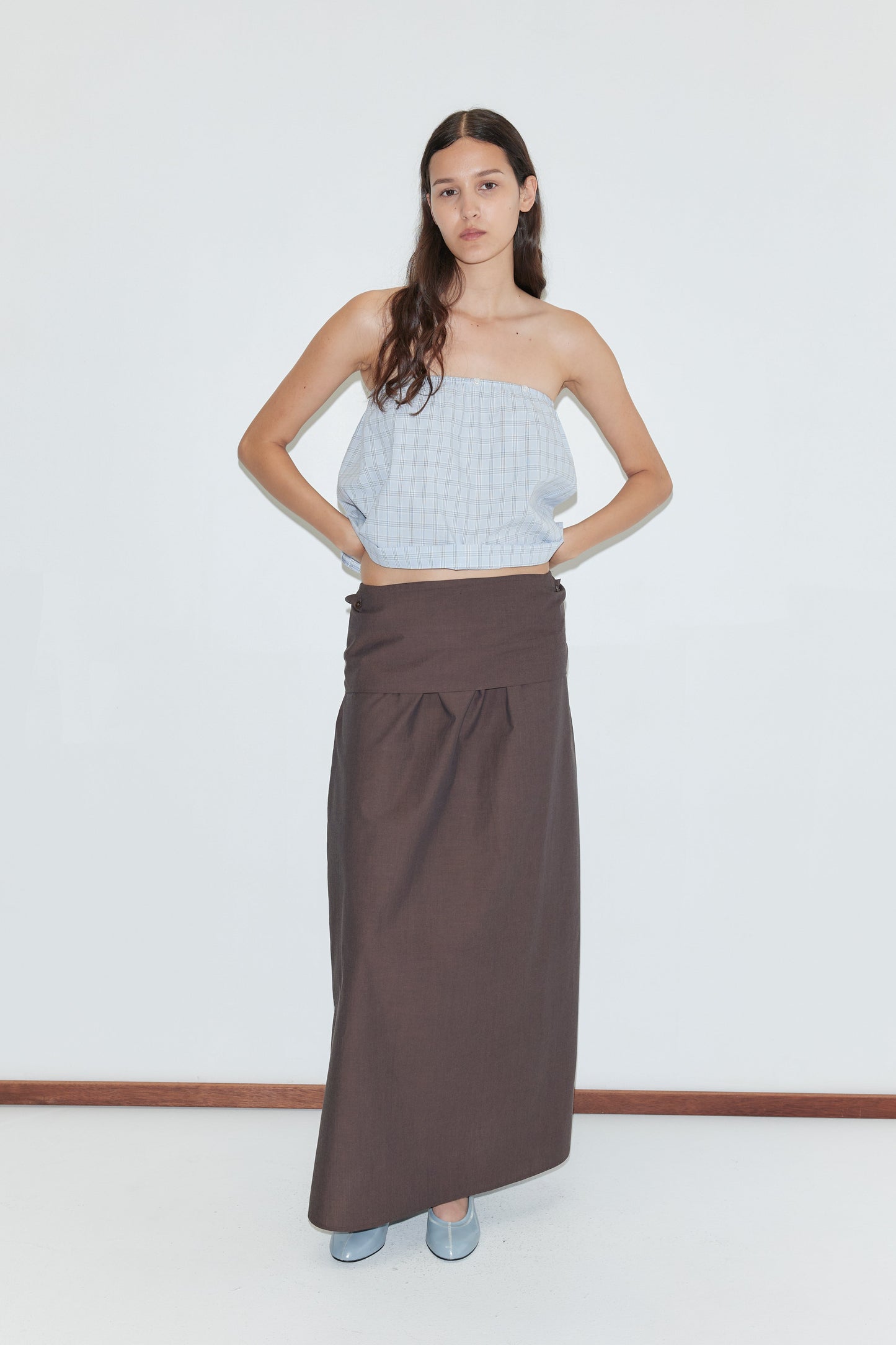 A woman stands against a white background in a strapless light blue checkered top, long brown skirt by Deiji Studios, and the Shirt Cuff Belt in Toast. She has long dark hair, blue shoes, and her hands rest on her hips above the chic belt.