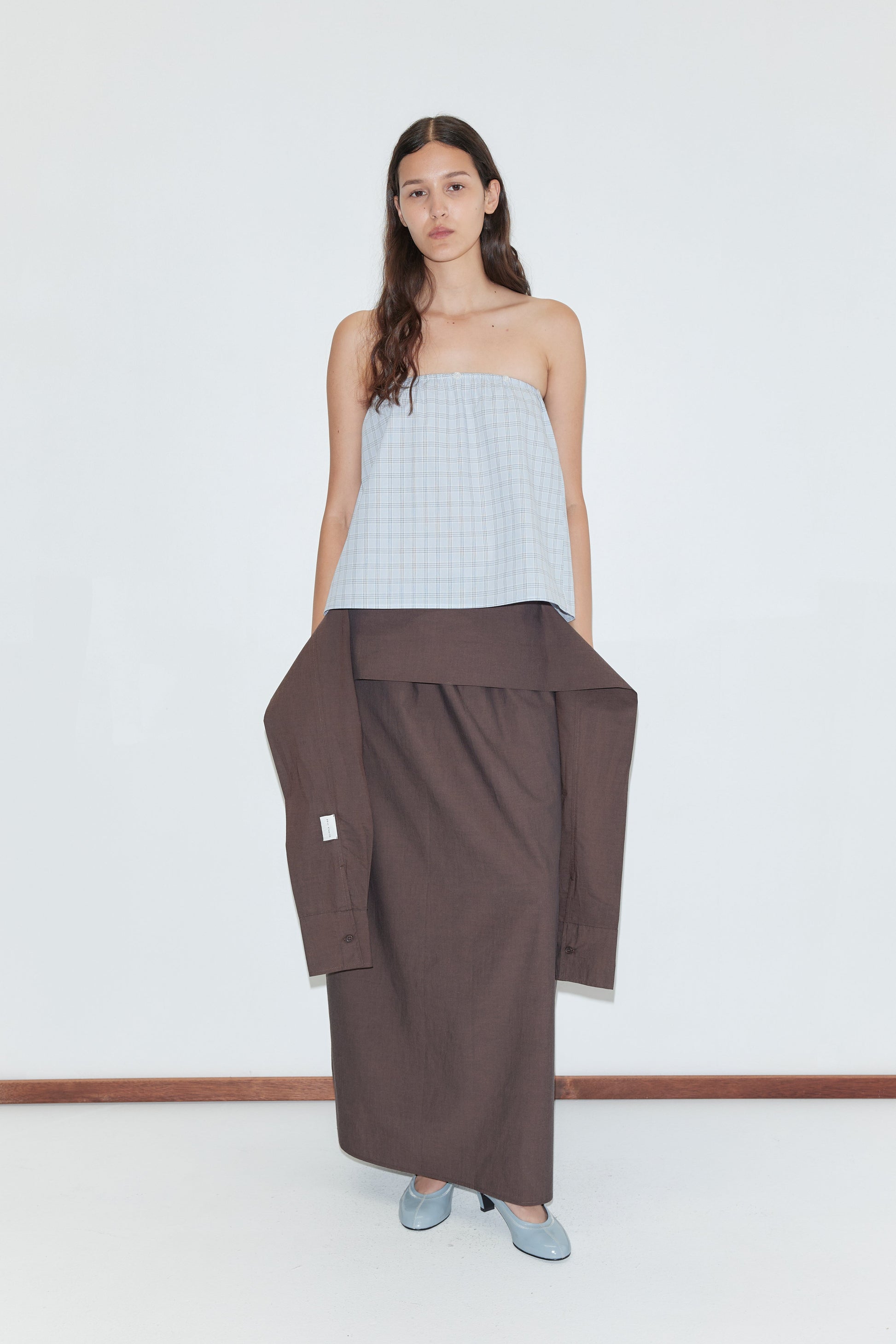 A woman stands against a plain white background wearing a blue plaid strapless top, a Deiji Studios long brown skirt, the Shirt Cuff Belt - Toast around her waist, and light blue flat shoes.