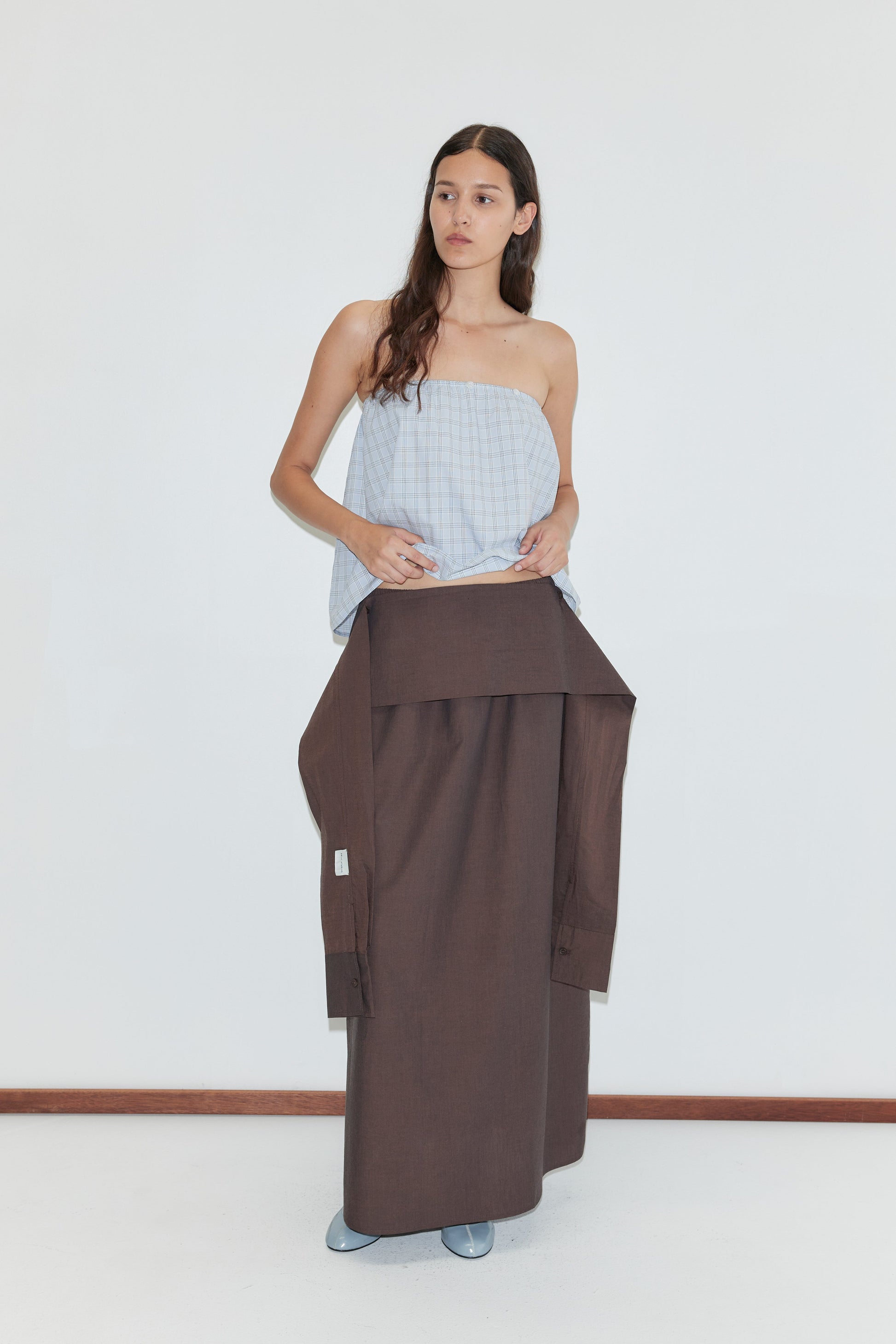 A woman with long brown hair stands against a white background, wearing the Way Skirt - Toast, a full-length brown skirt with an attached shirt detail at the waist, paired with a light blue strapless top and light blue shoes.
