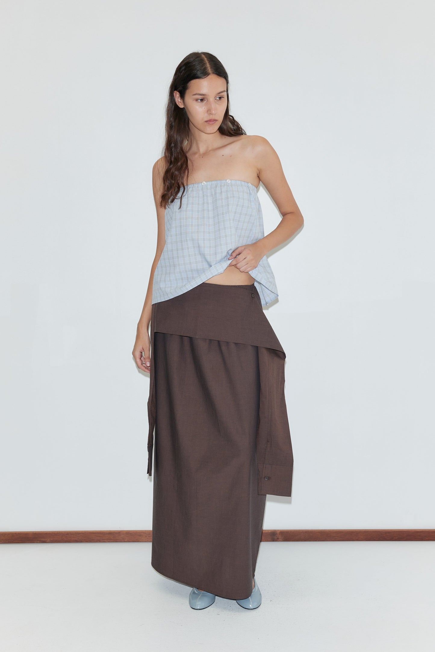 A woman with long brown hair wears a strapless, light blue checkered top and a long brown skirt featuring the Shirt Cuff Belt in Toast from Deiji Studios, standing against a plain white background and looking to the side.