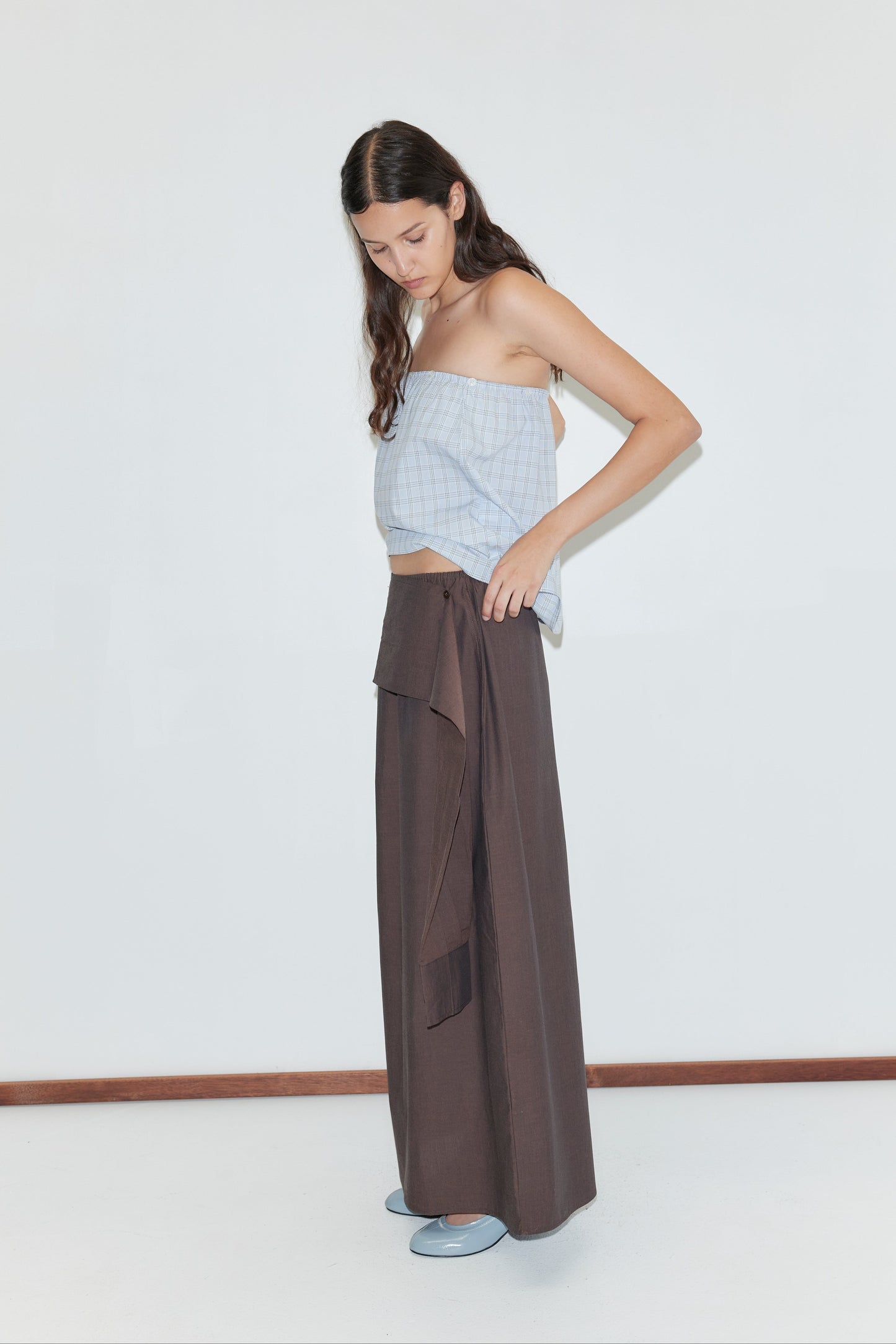 A woman with long brown hair indoors adjusts the Shirt Cuff Belt - Toast on her loose, dark brown wide-leg pants. She wears a light blue sleeveless checkered top and blue shoes, standing against a plain white background.