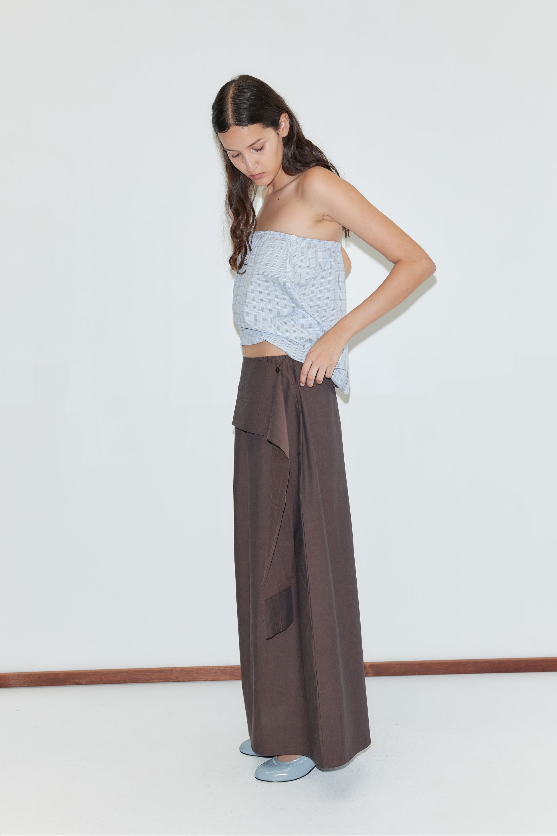 A woman with long brown hair indoors adjusts the Shirt Cuff Belt - Toast on her loose, dark brown wide-leg pants. She wears a light blue sleeveless checkered top and blue shoes, standing against a plain white background.