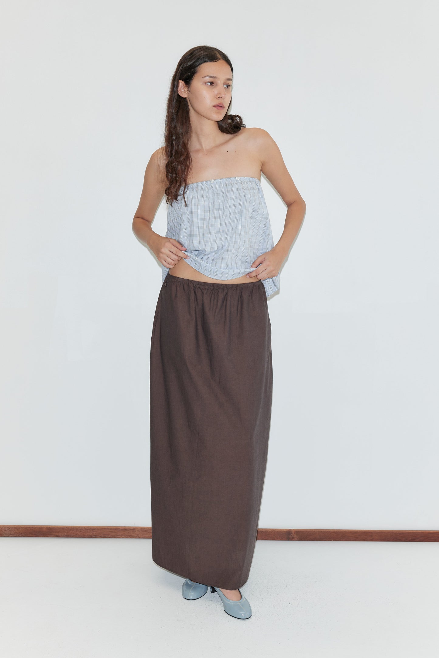 A woman with long, wavy hair stands indoors against a plain white background wearing the Way Skirt in Toast, a strapless light blue plaid top, and blue flat shoes. She looks to the side.