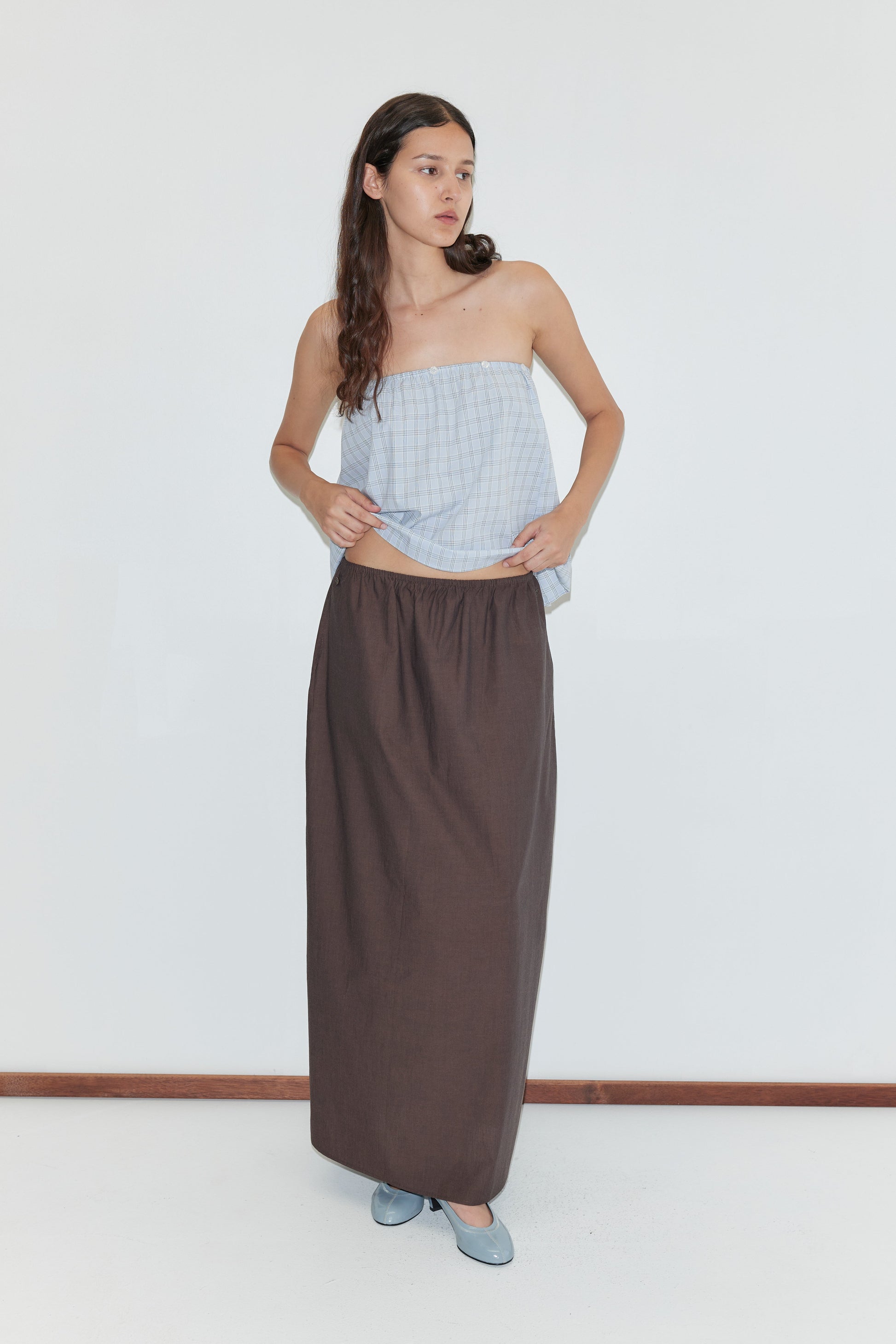 A woman with long, wavy hair stands indoors against a plain white background wearing the Way Skirt in Toast, a strapless light blue plaid top, and blue flat shoes. She looks to the side.