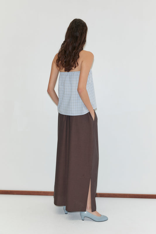 A woman with long brown hair stands facing away, wearing a Deiji Studios light blue checkered sleeveless top and the Way Skirt - Toast, an organic cotton full-length skirt with a side slit, her hands in the skirt pockets and light blue shoes.