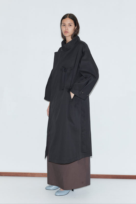 A person stands against a plain white wall wearing the Longline Jacket - Black from Deiji Studios over a brown skirt and light gray shoes. Their hair is parted in the middle and loose, with a calm, neutral expression.