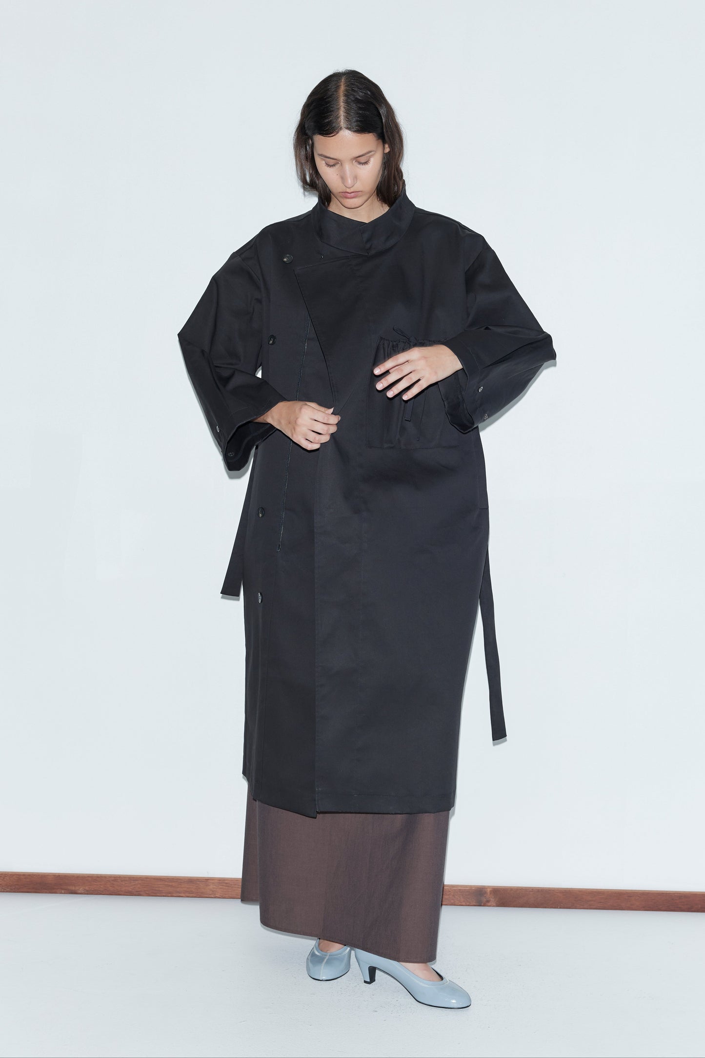 A person stands against a white background wearing the Longline Jacket - Black from deiji studios over a brown skirt and light blue heels, looking down while adjusting the jacket with one hand.