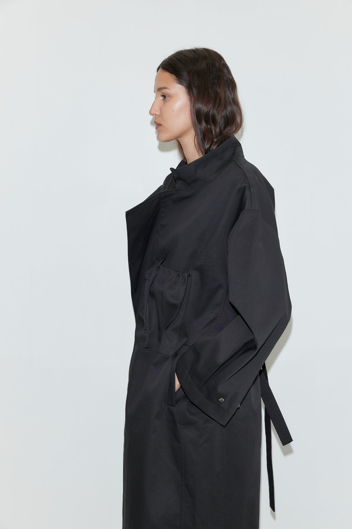 A person with wavy dark hair stands in profile against a plain white background, wearing the Longline Jacket - Black from Deiji Studios, hands tucked casually into the pockets.