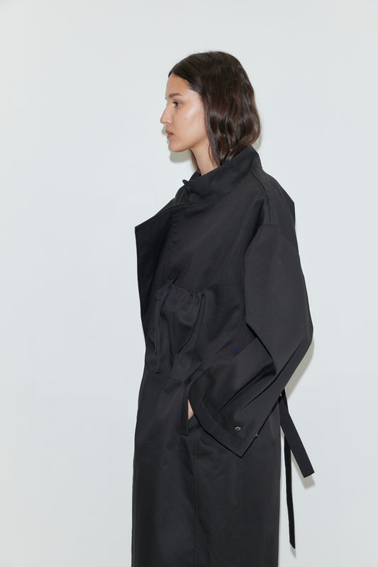 A person with wavy dark hair stands in profile against a plain white background, wearing the Longline Jacket - Black from Deiji Studios, hands tucked casually into the pockets.